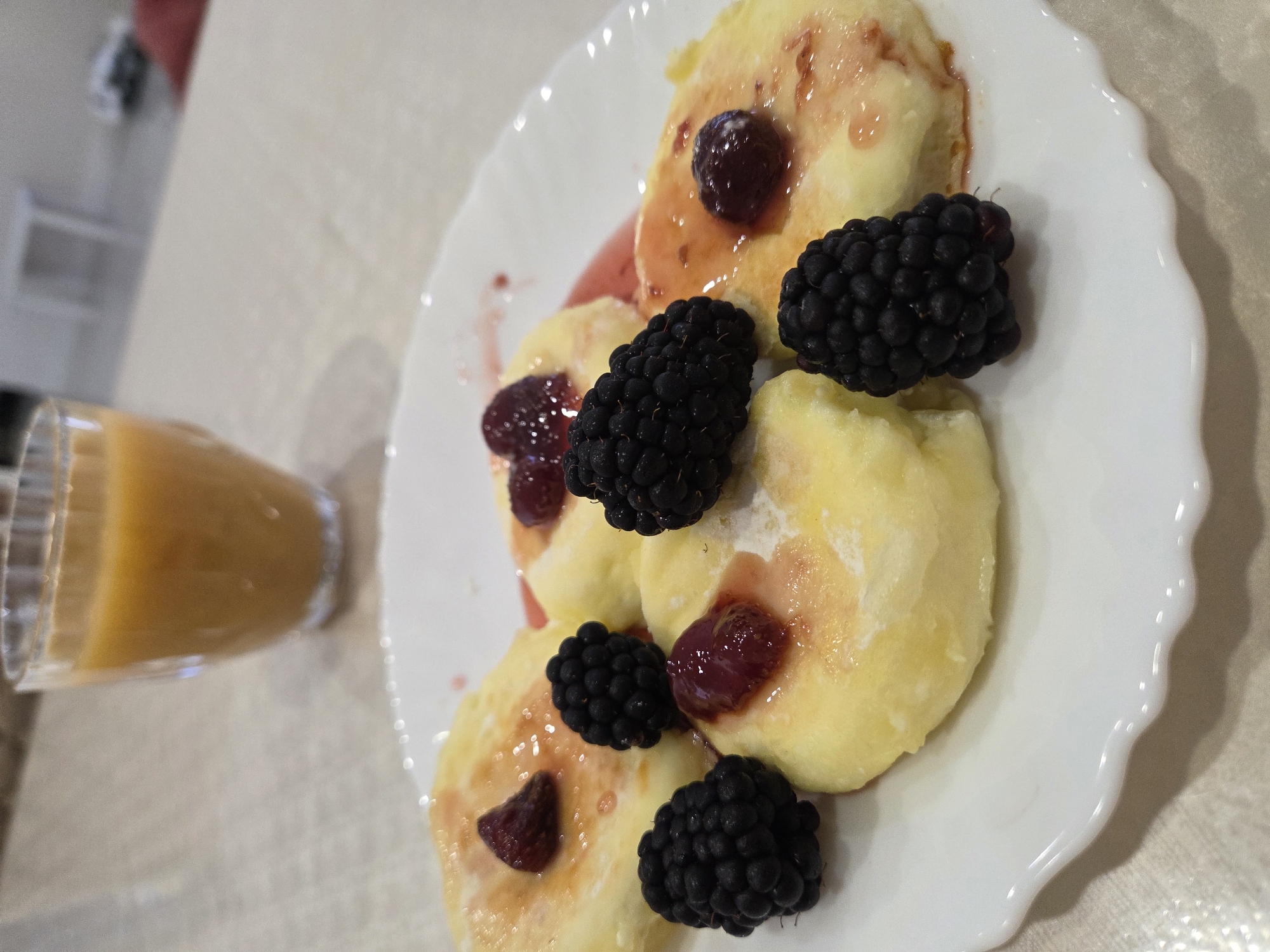 A close-up, eye-level shot shows a white, scalloped-edge plate with several syrniki (Russian cheese pancakes) arranged on it. The syrniki are golden yellow, slightly irregular in shape, and topped with a pinkish syrup and small, dark red berries, likely raspberries or cranberries. Several fresh blackberries are placed around the syrniki, with one on top of a syrnik. In the background, a clear glass filled with orange juice or a similar beverage is visible, set against a light-colored, textured surface that appears to be a tablecloth or placemat. The overall setting is likely a home or cafe. There are no people or animals in the image. The lighting is soft, and the focus is primarily on the food. There is no discernible context linking this scene to Ulyanovsk, Russia; it is a generic image of food.
