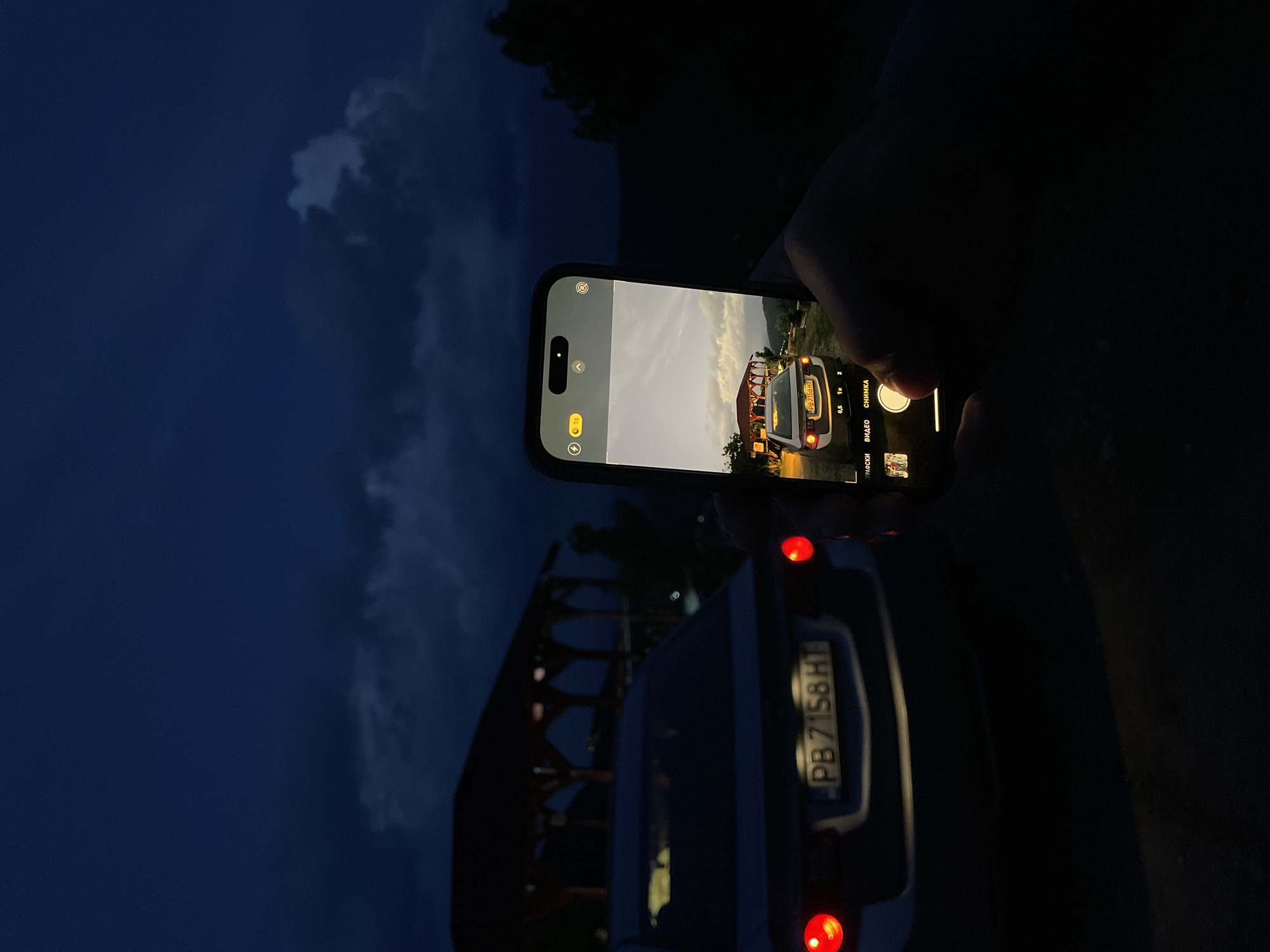 A person holds a smartphone and takes a photo of a parked car with a surfboard carrier on top. The car has a Bulgarian license plate that reads "PB 7158 HT". The smartphone's screen shows a brighter image of the car and its surroundings, potentially reflecting an earlier time of day. The background shows a dark sky, possibly at dusk or dawn. The location is likely in Pechinska, Bulgaria.