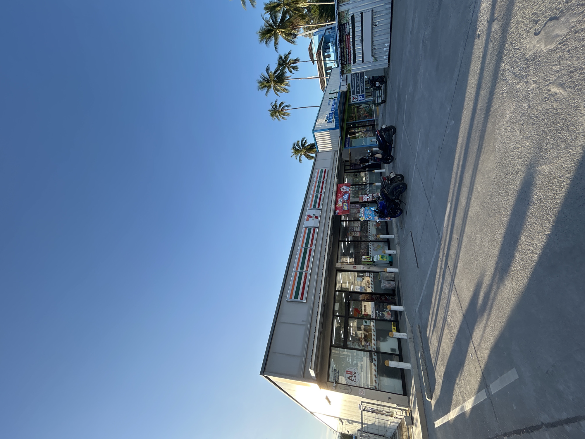 The image depicts a commercial storefront, likely a convenience store, in Baan Hua Thanon, Thailand. The facade features the prominent "7-Eleven" logo. In front of the store, several motorcycles are parked on a paved surface. The area is illuminated by natural sunlight, casting long shadows across the ground, suggesting it is either early morning or late afternoon. The sky is clear and blue, with a few palm trees visible in the background, indicating a tropical environment. The building appears to be a single-story structure with a modern, functional design.