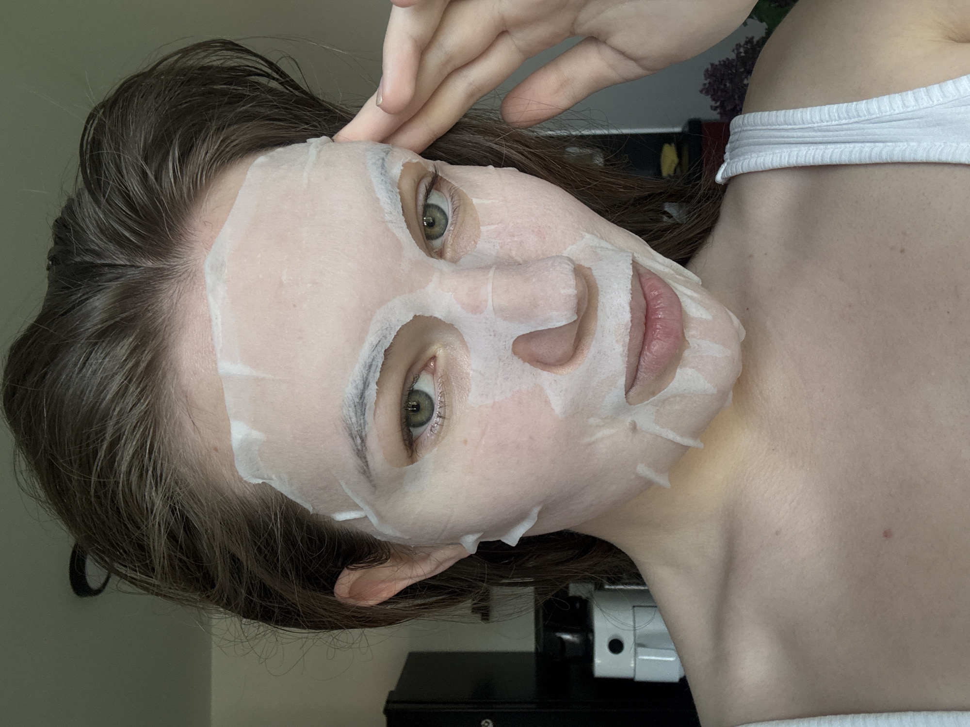 A person with a facial mask is looking at the camera, tilting their head. The person has green eyes and dark brown hair. They are wearing a white top. The scene appears to be indoors. The location cannot be determined.