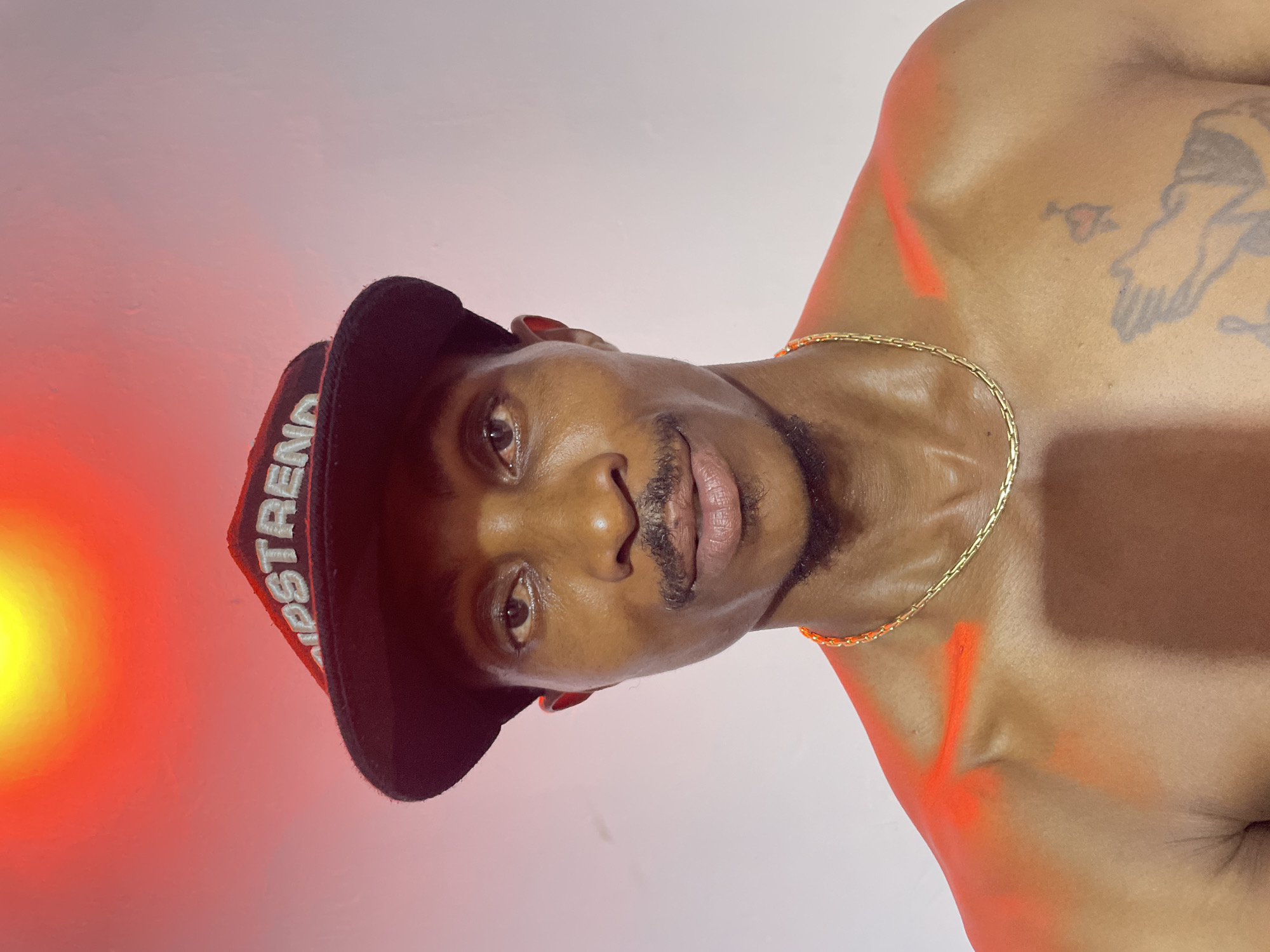 A head-and-shoulders, medium shot portrait of a Black man wearing a black baseball cap with "UPSTREND" embroidered in white lettering. He has a thin mustache and goatee. He is wearing a gold chain necklace. His bare shoulders are visible and are partially illuminated by red and orange light. There is an orange mark on his right shoulder. A tattoo of praying hands is visible on his chest. The background is a white wall with a bright yellow and red light source in the upper left corner. There is no visible indication of the specific location being Adankolo, Nigeria.