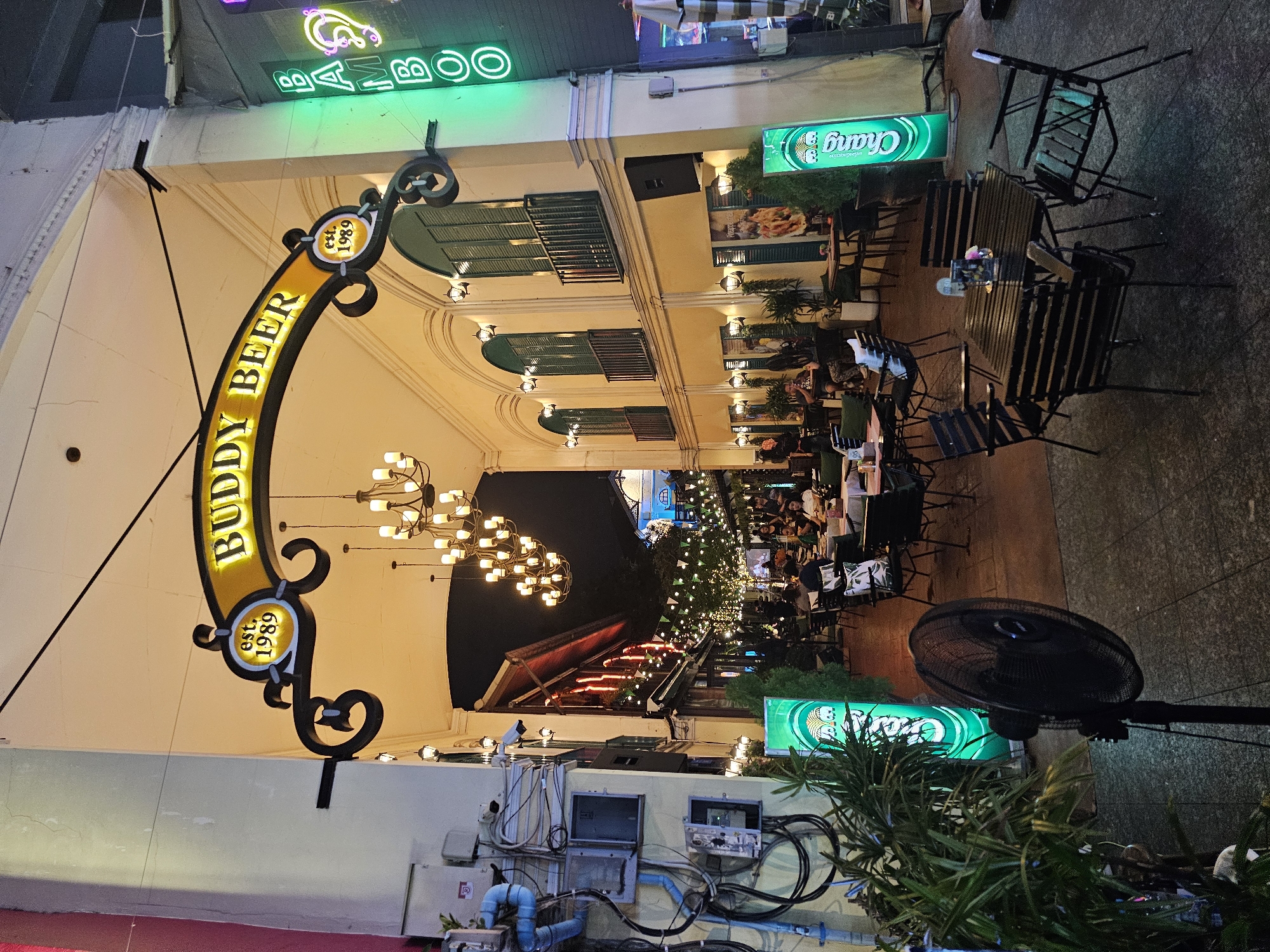 The image depicts an outdoor dining area at night, likely a restaurant or bar, located in Bangkok, Thailand. A large, illuminated yellow archway sign reads "BUDDY BEER" with "est. 1989" prominently displayed. To the right of the archway, a vertical green neon sign spells out "BAMBOO". The scene is set under an arched building facade with multiple windows and balconies. Numerous tables and chairs are arranged on a tiled floor, with some tables occupied by patrons. String lights and ornate chandeliers illuminate the area. Several green illuminated signs, likely for the beer brand "Chang," are visible throughout the space. A black oscillating fan stands in the foreground, and dense greenery is present on the left side of the image. Electrical conduits and boxes are mounted on the wall to the left.