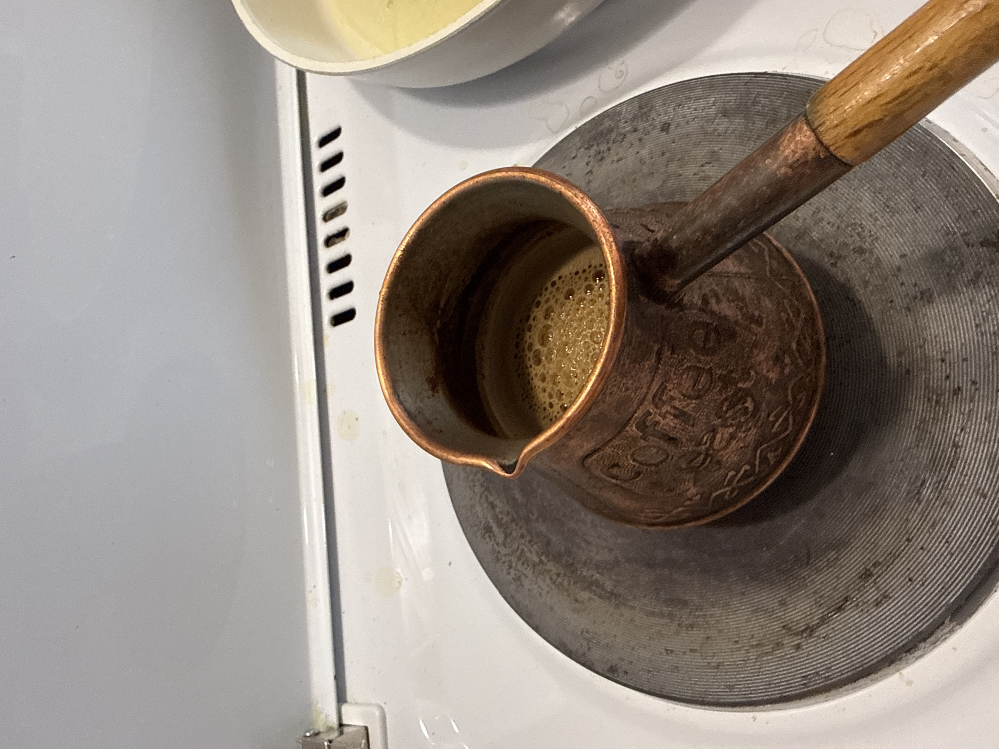 The image presents a high-angle, close-up view of a copper cezve (Turkish coffee pot) actively brewing coffee on an electric stovetop. The cezve, featuring a long wooden handle and a spout, is positioned on a round, metallic electric burner that shows signs of use and heat discoloration. Inside the cezve, dark brown coffee is visible, topped with a thick, light brown layer of frothy foam and small bubbles, indicating it is hot and freshly prepared.
To the left of the burner, a white surface of the stovetop is visible, alongside a vertical panel with evenly spaced, dark ventilation slits or a grate. In the upper left corner, a portion of a white bowl or pot containing a pale yellow substance, possibly a liquid or batter, is partially in view.
The copper body of the cezve is aged and exhibits a visible patina. Faintly embossed in decorative script on its side is the word "COFFEE". The overall setting suggests a domestic kitchen environment in Tobolsk, Russia, with the focus on the preparation of a hot beverage. The lighting is diffused, typical of an indoor setting, and no specific time of day can be determined.