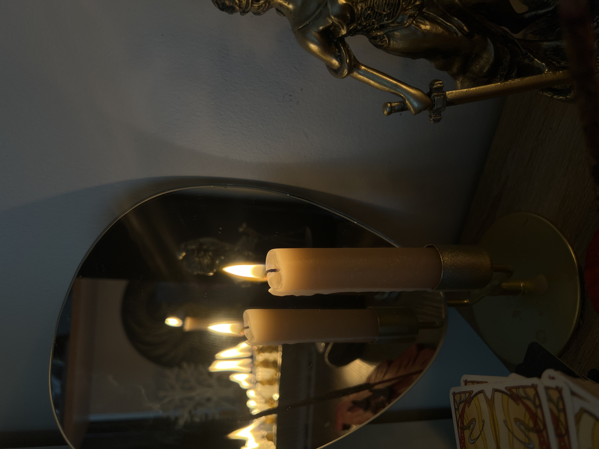A dimly lit indoor scene features two lit candles in brass holders on a wooden surface. The candles are reflected in a large, heart-shaped mirror positioned behind them. The mirror's reflection shows multiple candle flames, some appearing as a blurred line of light. To the right of the candles, a golden statue of Lady Justice, holding a sword and scales, stands partially in view. In the foreground, a stack of tarot cards is visible on the left. The background is a textured wall. The lighting suggests it is nighttime, and the overall atmosphere is mysterious. There is no indication of the location being in Tychy, Poland.
