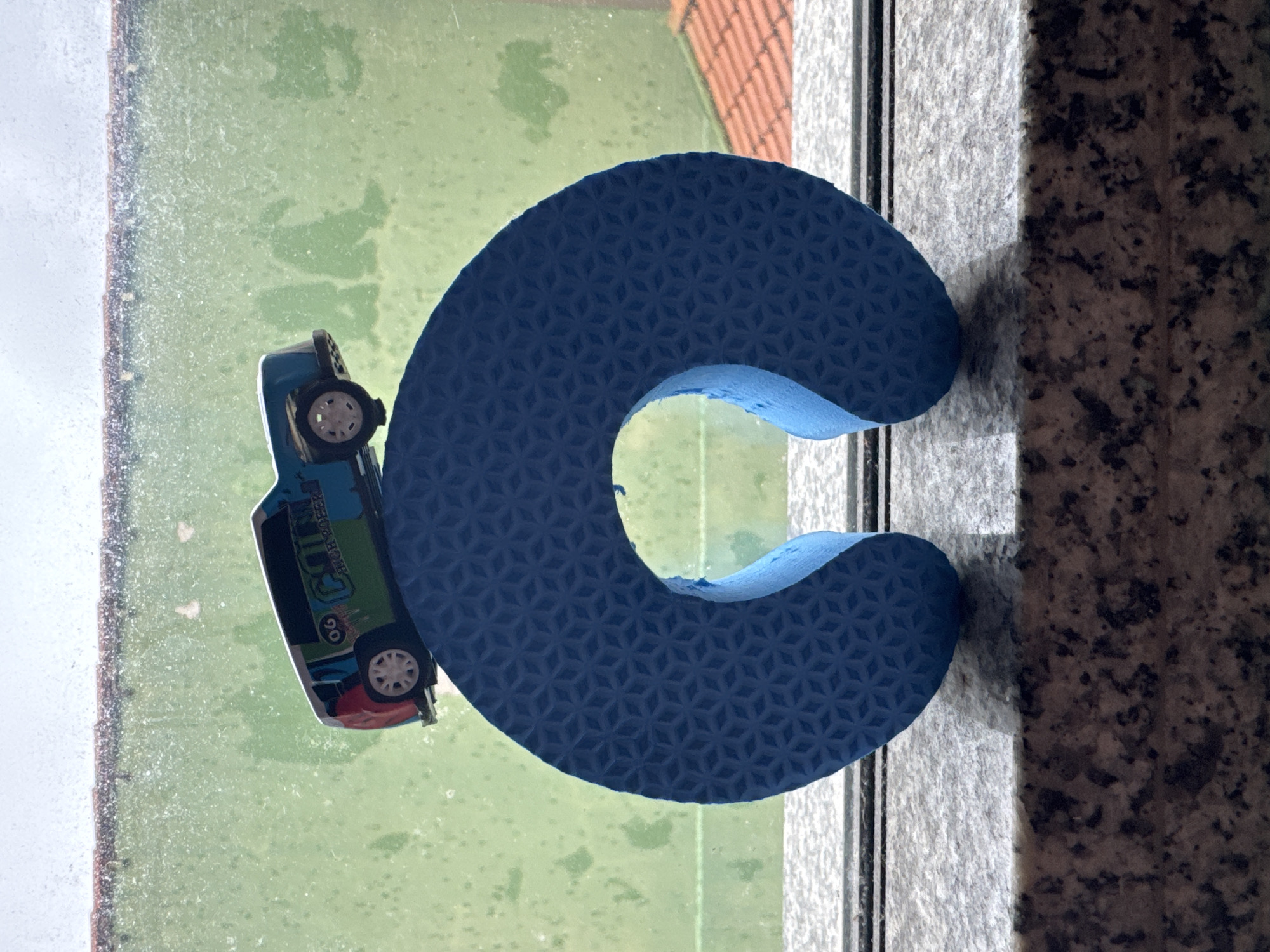 A blue neck pillow, embossed with a geometric pattern, rests on a speckled granite surface. Perched atop the pillow is a small, toy van with a multi-colored design and the number "06" visible on its side. The background consists of a green, rain-streaked window, hinting at an outdoor environment. Beyond the window, a tiled roof and some foliage are discernible. The scene is indoors, likely within an apartment or house, positioned near a window. The image does not provide any specific indicators of location in Niterói, Brazil, or any people.