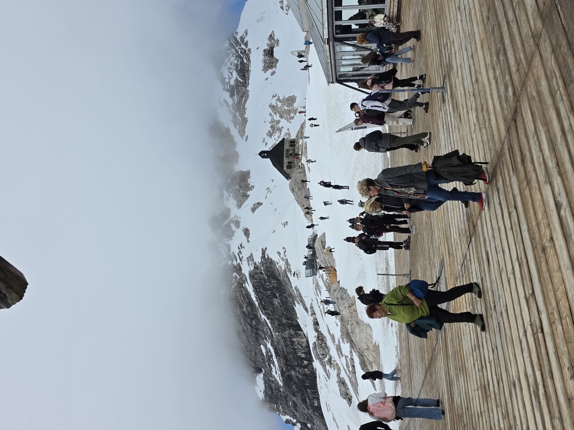The image captures a high-altitude scene on a snowy mountainside, likely at the Zugspitze, Germany's highest peak, near Garmisch-Partenkirchen. A group of tourists is walking on a wooden platform in the foreground. The platform is constructed with wide, light-colored wooden planks laid horizontally, creating a textured surface. The background features a dramatic mountain landscape covered in snow and rocky outcrops, with a small, dark-roofed building, possibly a chapel, perched on a rocky ledge. Clouds partially obscure the upper reaches of the mountain, adding a sense of vastness and dramatic weather. Tourists are scattered across the snowy slopes and the platform, engaged in various activities such as walking, observing the scenery, and congregating near a modern glass-enclosed building to the right of the frame. The people are dressed in casual attire, with many wearing jackets and carrying backpacks, suitable for a cool mountain environment. The overall scene depicts a popular tourist destination offering panoramic views of the alpine scenery.