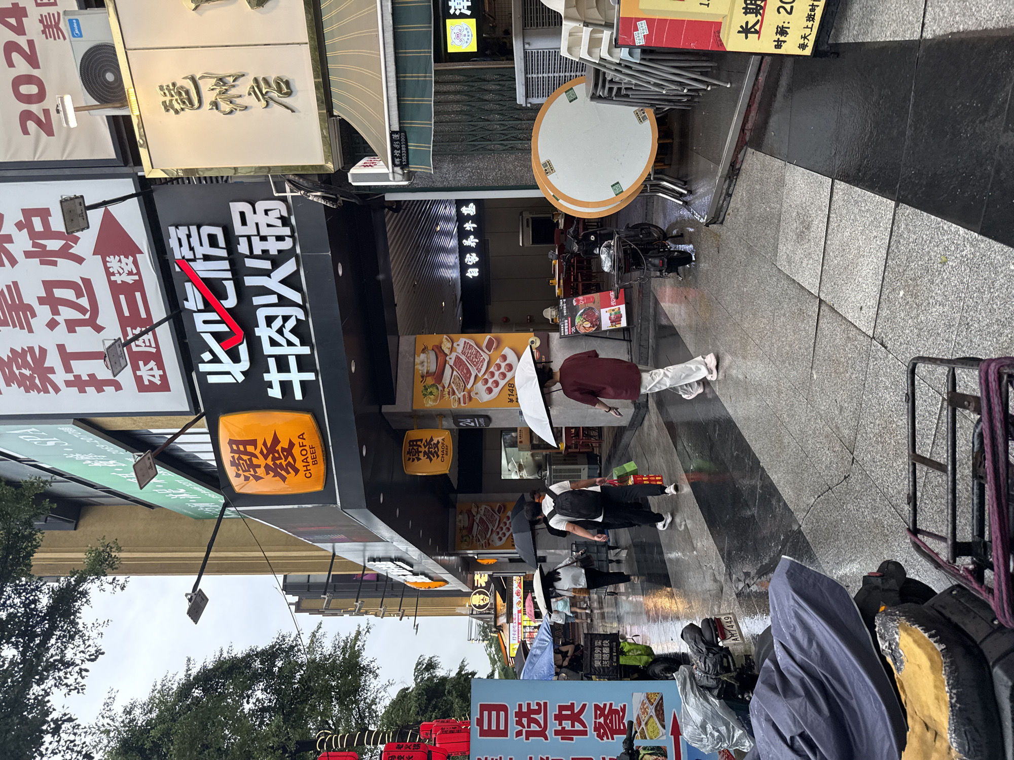 This is a photograph taken from a street-level perspective, showing a bustling commercial area in Haizhu District, China. The image is oriented vertically, with the bottom of the scene appearing at the top of the frame. The weather appears to be rainy, as evidenced by the wet pavement and several people holding umbrellas. The sky is overcast, suggesting it is daytime, likely late morning or afternoon.
The scene is dominated by shopfronts with prominent signage in Chinese, advertising restaurants and other businesses. One large sign displays "必吃情 牛肉火锅" (Must-eat Beef Hot Pot), with a red arrow pointing upwards. Another sign reads "潮發 BEEF" in both Chinese and English. There are also signs related to food delivery services, like "美团外卖" (Meituan Waimai). Objects visible include stacked chairs and tables outside a restaurant, a motorbike, and various shopping carts and bags, some covered with tarps.
Several people are present in the image, most of whom appear to be walking or standing on the sidewalk. Some are using umbrellas to shield themselves from the rain. The overall mood seems to be one of daily commerce continuing despite the weather. The presence of trees on the left side of the image suggests a mix of urban and greener spaces. The time of day is indicated by text on a sign that reads "时新:20 每天上班时," suggesting operating hours.