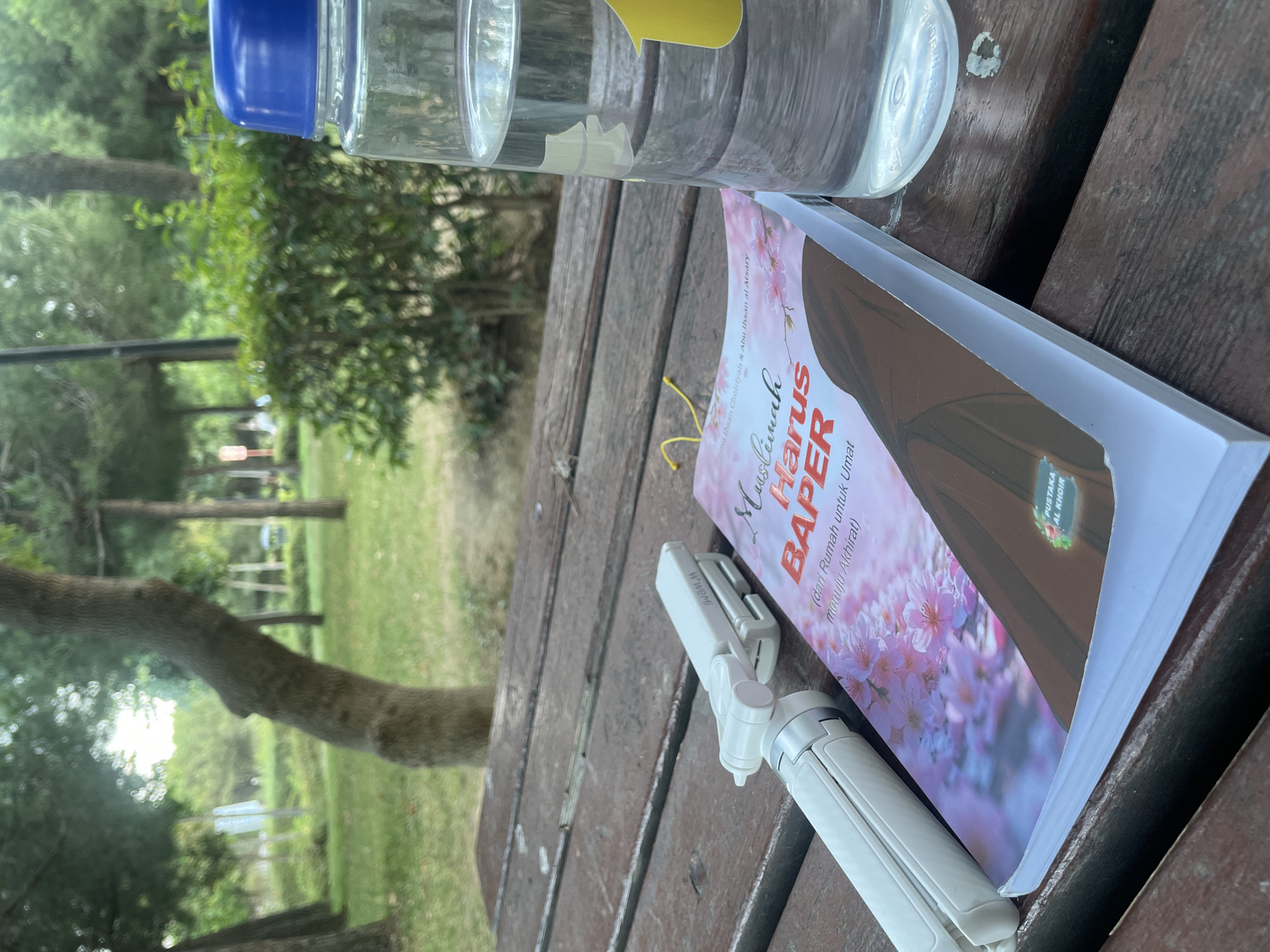 A close-up, eye-level shot shows a wooden picnic table in a park. On the table, a book with a cover featuring pink flowers and a woman in a hijab is open, next to a white tripod and a clear plastic water bottle filled with water. The background is blurred, revealing trees and green foliage, suggesting an outdoor setting. The overall scene evokes a sense of calm and leisure, likely in a park within Başakşehir, Turkey.