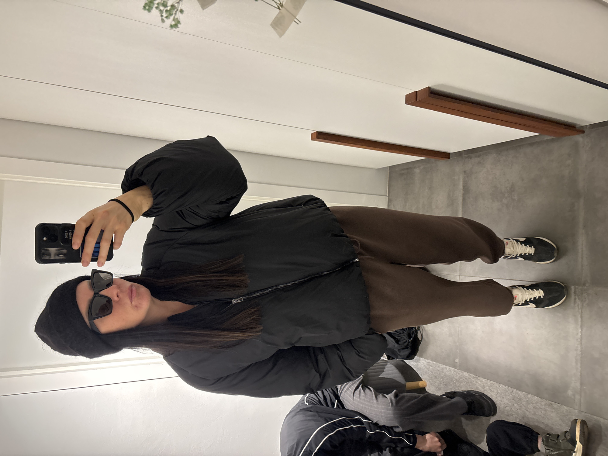 A person wearing a black beanie, sunglasses, a black puffer jacket, and brown sweatpants is taking a selfie in a room with white walls and grey flooring. Visible in the reflection is another person wearing grey striped pants and black shoes. A plant decoration is visible on the wall above. Wooden bars are attached to the ceiling. The location is a store in Chaikovsky, Russia.