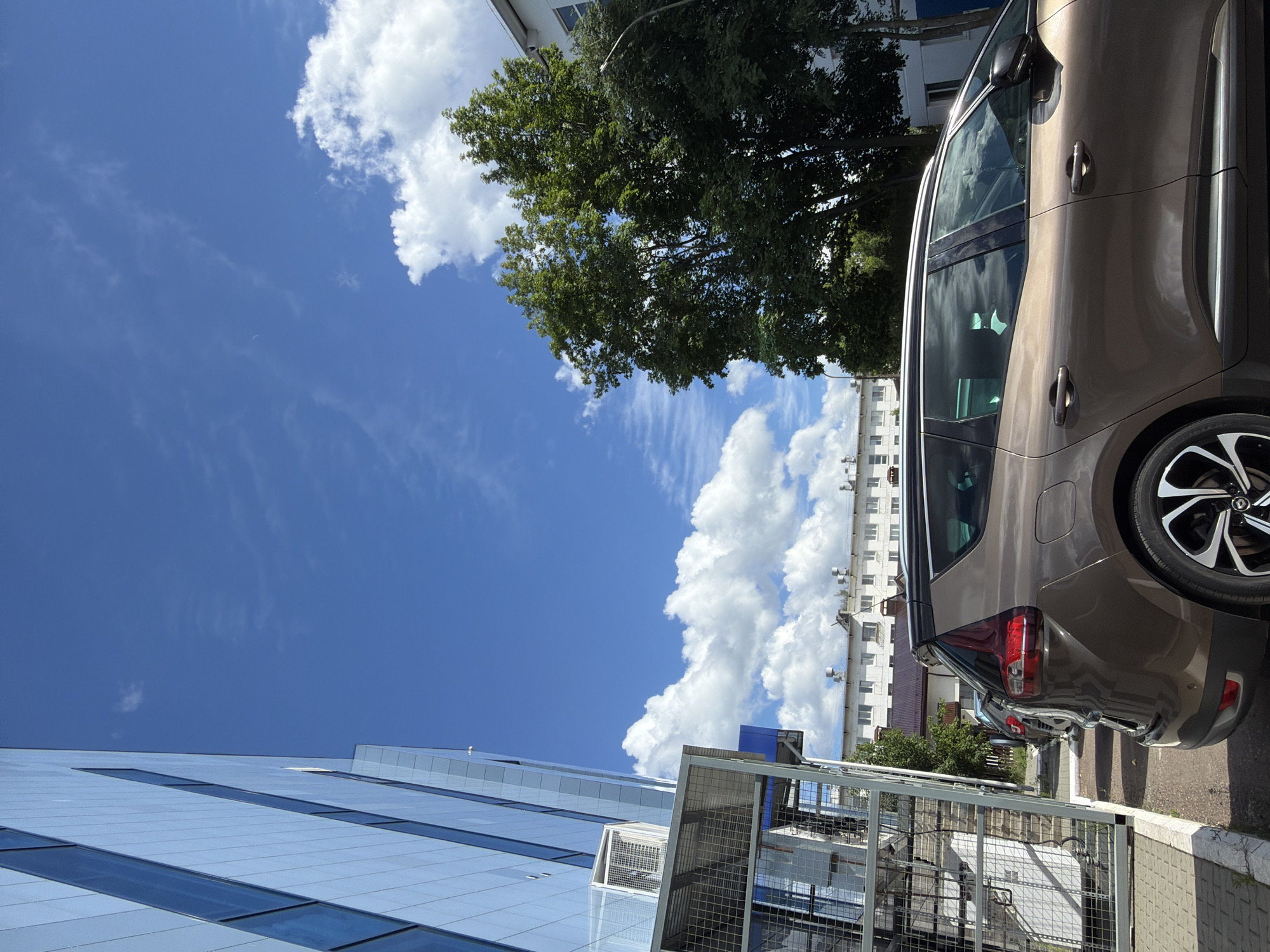 This is a vertical, eye-level outdoor photograph taken on a sunny day with fluffy white clouds scattered across a bright blue sky. The scene depicts a street or parking area with modern architecture and a car.
In the foreground, a light brown car is parked, its side prominently featured. The car's windows reflect the sky and clouds. Behind the car, a fence with a metal mesh construction is visible, suggesting a secured area or property boundary. Further in the background, a multi-story building with a light-colored facade and numerous windows can be seen. Adjacent to this building, lush green trees with dense foliage add a natural element to the urban landscape. Another, more modern building with a predominantly glass facade and horizontal blue-tinted windows is on the left side of the frame. The overall setting appears to be a commercial or residential area in Chaikovsky, Russia.
The lighting and clear sky indicate it is daytime, likely mid-morning or afternoon. The weather appears pleasant and sunny, conducive to outdoor activities. No people are visible in the frame, and no text is discernible. The image conveys a sense of everyday life in a modern urban environment, characterized by a mix of architecture, vehicles, and greenery.