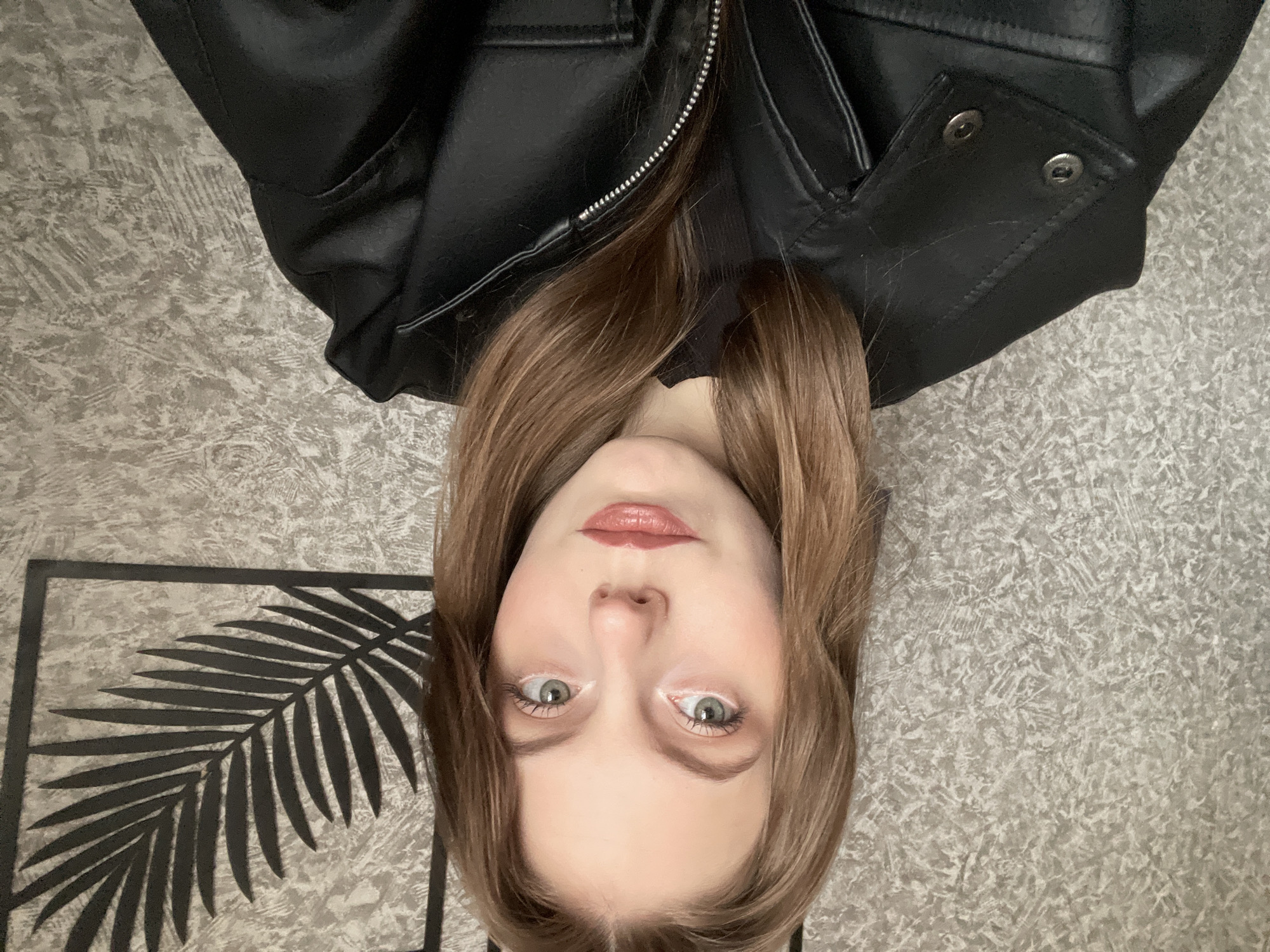 A young woman is captured in a close-up self-portrait, lying down and looking directly at the camera. Her face is positioned towards the bottom center of the frame, with her long, straight brown hair fanned out around her head and extending upwards. She has fair skin, green/hazel eyes accentuated with white eyeliner on her lower waterline, and full lips colored with light pink lipstick. Her expression is neutral and engaging.
She is wearing a black leather jacket, visible across the upper portion of the image, with a prominent silver zipper down the front and silver snap buttons on a lapel or collar detail on the left side. Underneath the jacket, a black top is faintly visible.
The background consists of a light grey and beige mottled or textured surface, possibly a carpet, bedspread, or wall covering, giving an impression of an indoor setting. To the lower left of her head, a dark, stylized silhouette of a palm leaf framed by a rectangular border is visible on the textured surface, acting as a decorative element. No text is visible in the image. The image is taken in Фоки, Russia.