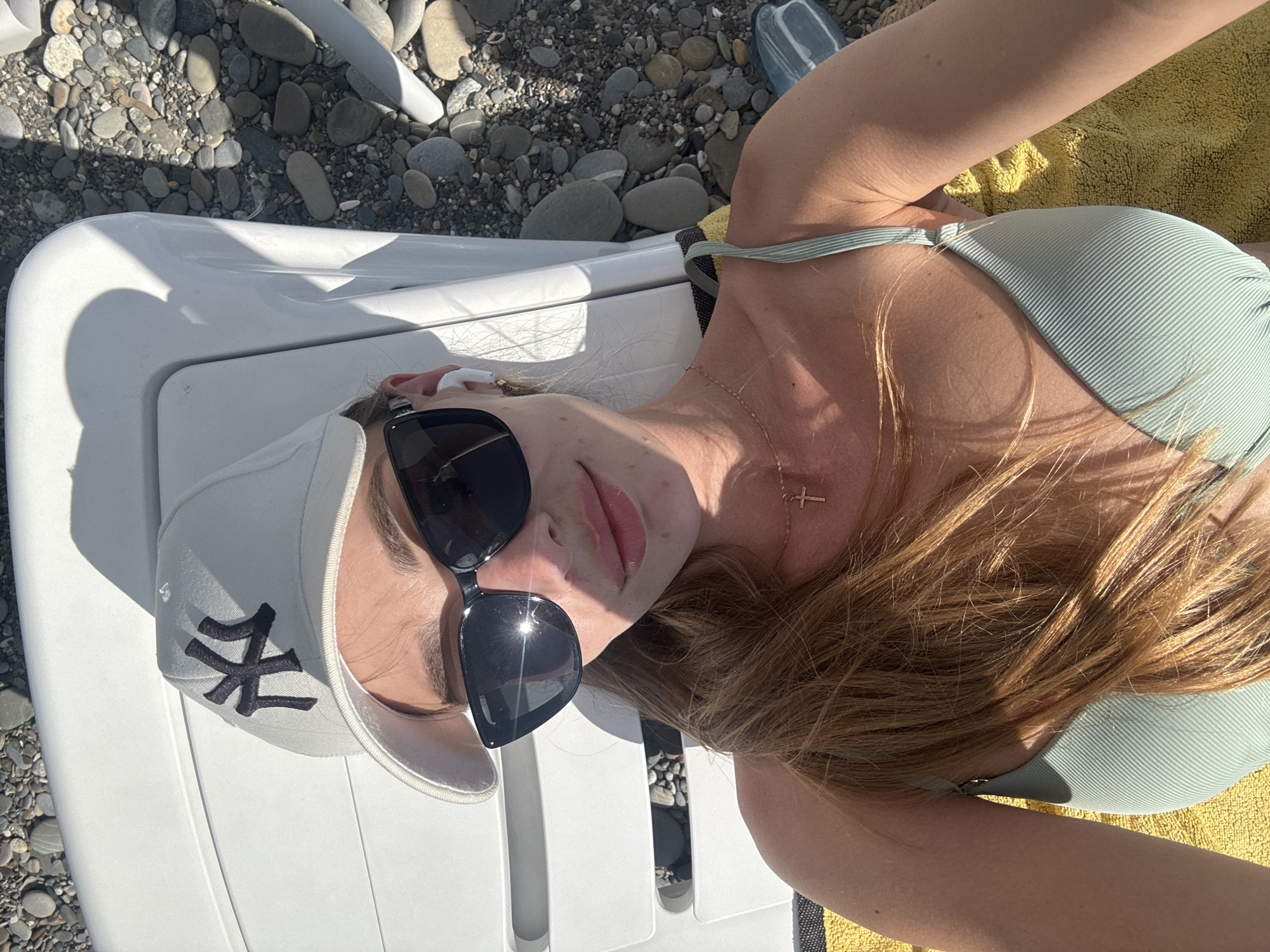 A young woman is lying on a white chaise lounge on a beach. She is wearing a white baseball cap with a black New York Yankees logo, black sunglasses, and a light green ribbed bikini. She has long, reddish-blonde hair and is wearing a delicate gold necklace with a cross pendant and white wireless earbuds. The beach is covered in pebbles, and the sea is not visible. The lighting suggests it is a sunny day. The specific location within Sochi, Russia, cannot be determined from the image.