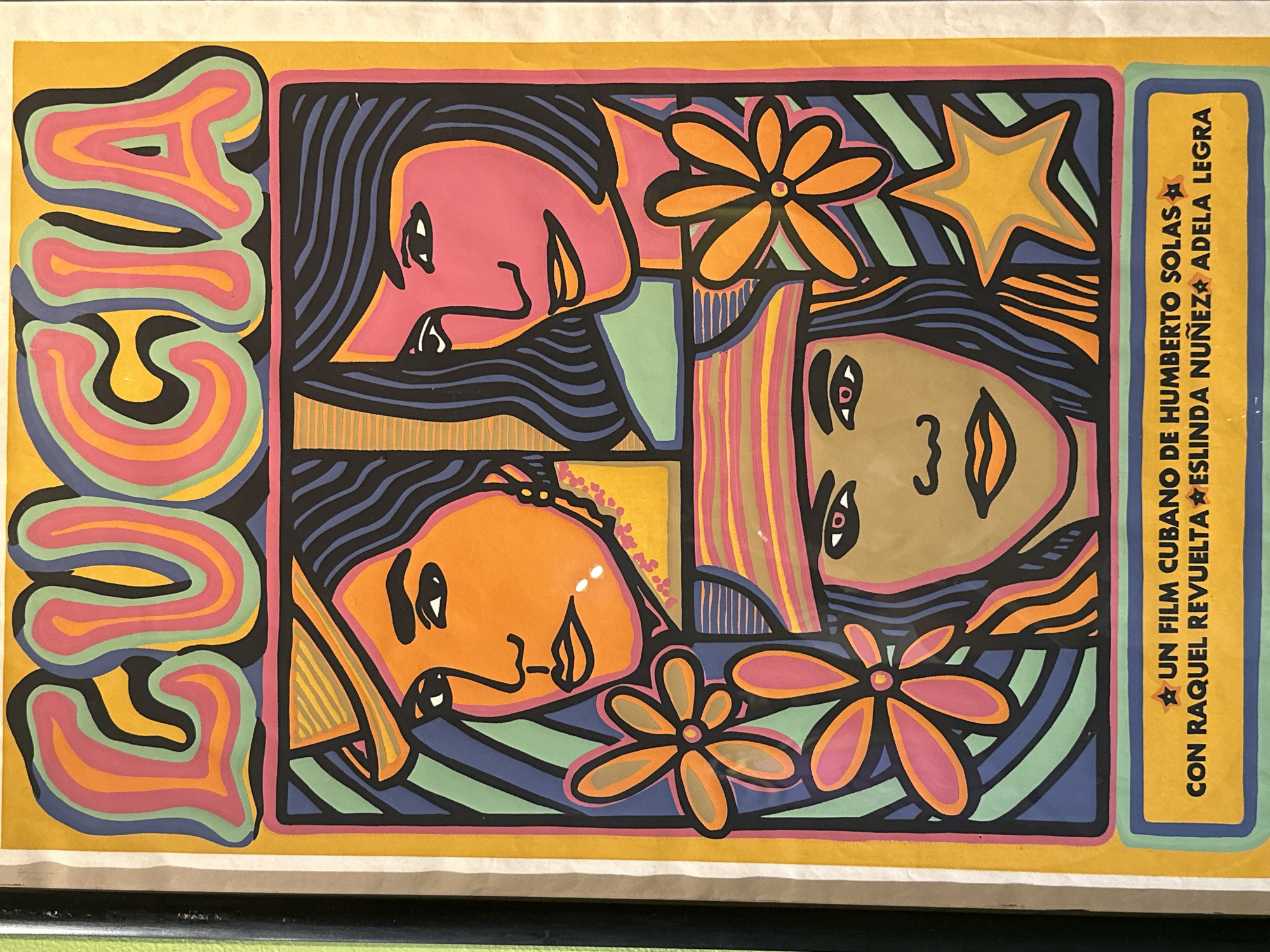 The image shows a poster with a psychedelic, pop art style. Vertically written on the left is the word "LUCIA" in wavy, multicolored outlines. To the right of the word is a square-ish panel filled with two pairs of faces oriented sideways and upside down. The faces are simplified and stylized with black hair. The faces are colored in pink, orange, and tan. Scattered around the faces are flower and star shapes. To the right of the artwork is a rectangular text block. The text reads "UN FILM CUBANO DE HUMBERTO SOLAS CON RAQUEL REVUELTA ESLINDA NUÑEZ ADELA LEGRA". There is one star before and one star after "UN FILM CUBANO DE HUMBERTO SOLAS." The poster is on a mustard yellow background.