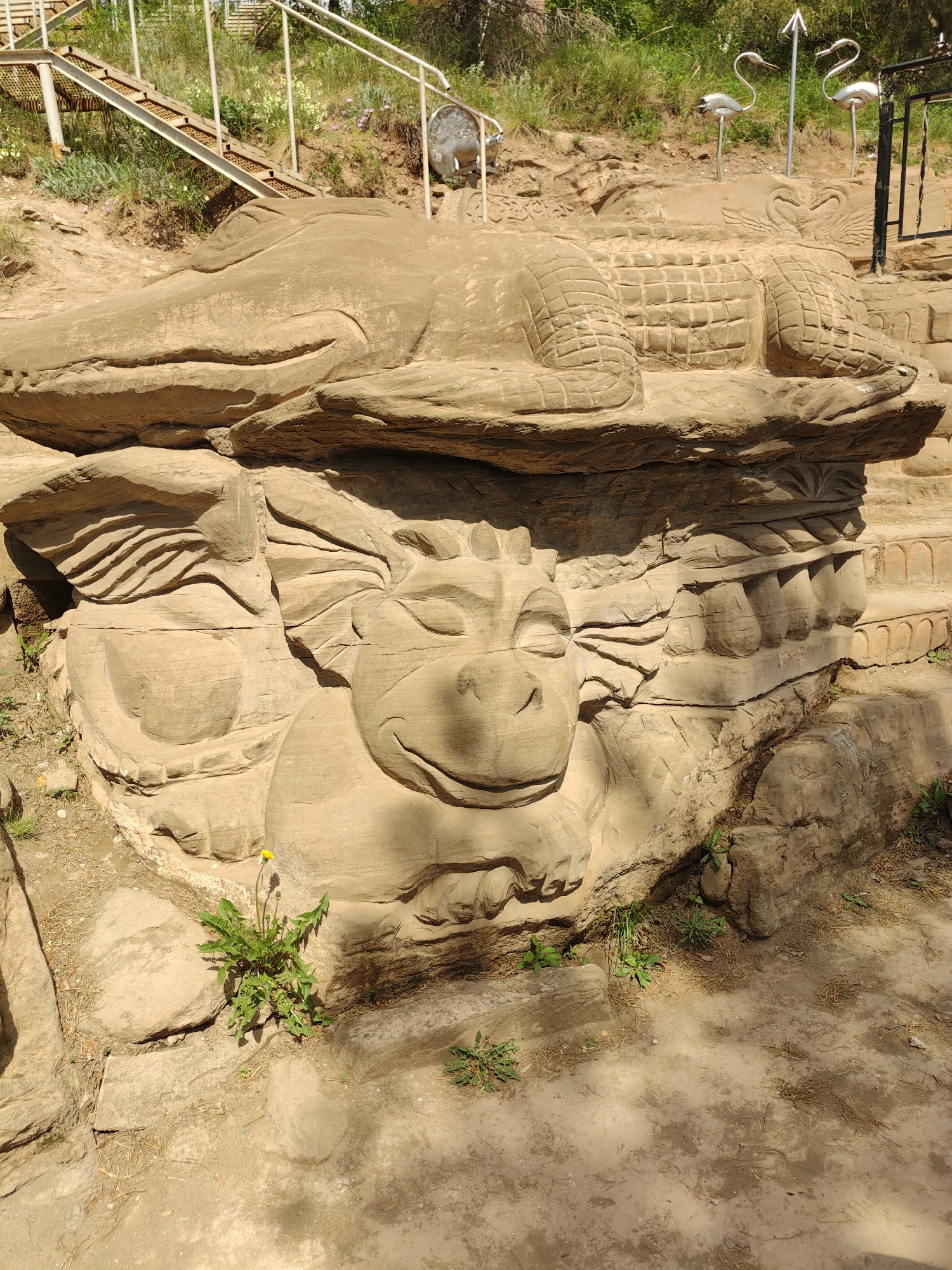 The image shows a sand sculpture on a rocky, possibly outdoor, setting. A crocodile-like figure with intricate details is sculpted on top. Below, a cartoonish, dragon-like face is carved into the rock. There are stairs with railings visible in the background, along with decorative metal bird figures. The scene is lit with natural sunlight, and some sparse vegetation grows around the base of the sculpture. The location is in the city of Unknown, Unknown.