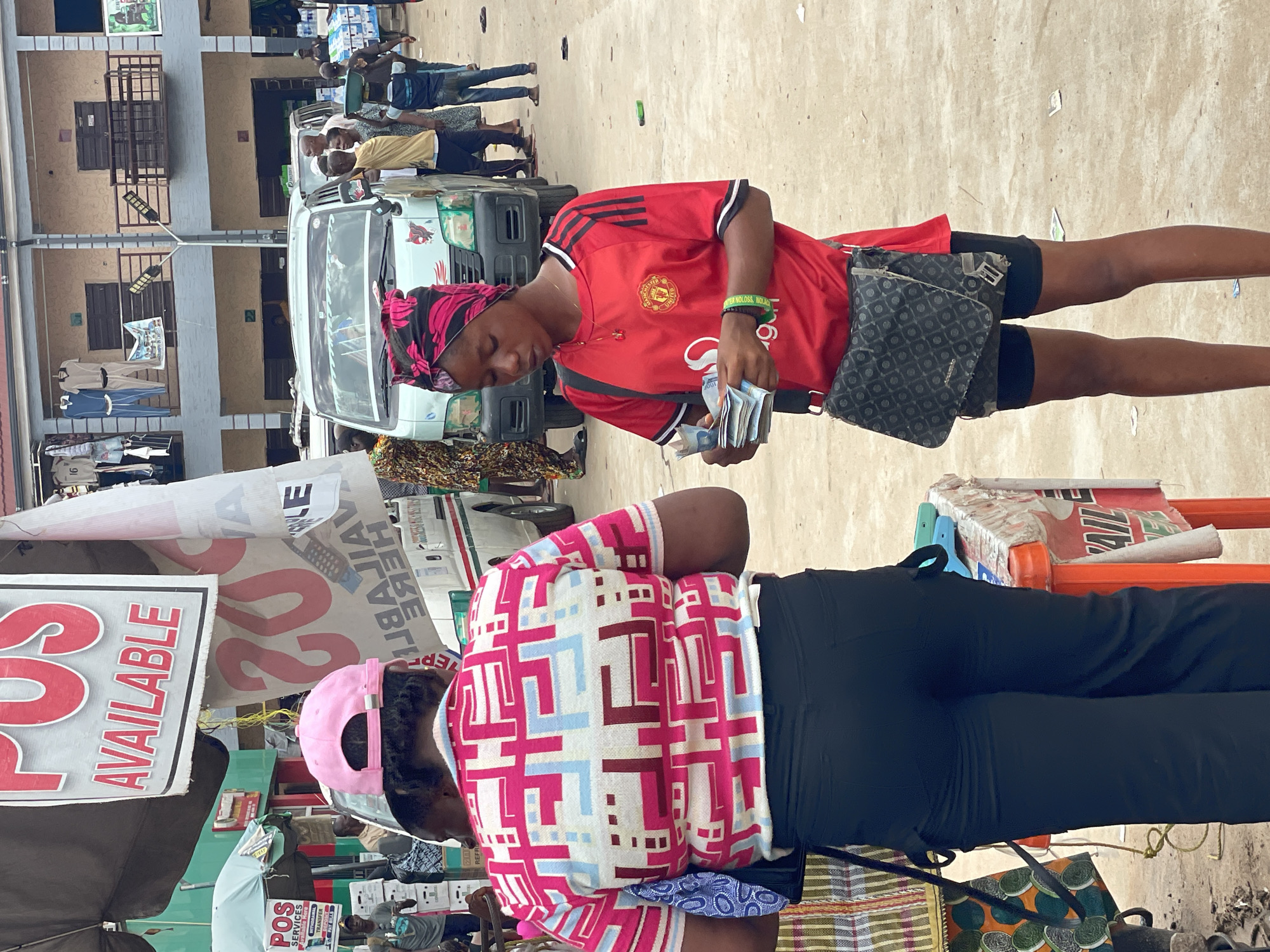 The image depicts a street scene in what appears to be a commercial area in Onitsha, Nigeria. A young woman wearing a red Manchester United jersey and a headscarf is counting Nigerian Naira notes. She has a crossbody bag. Beside her, another woman is wearing a pink hat and a colorful patterned shirt. They are positioned in front of a stall with a sign that says "POS SERVICES". Other signage includes "AVAILABLE" and "20 AVAILABLE HERE". In the background, there is a commercial vehicle and several people. The scene is crowded, suggesting a busy commercial environment.