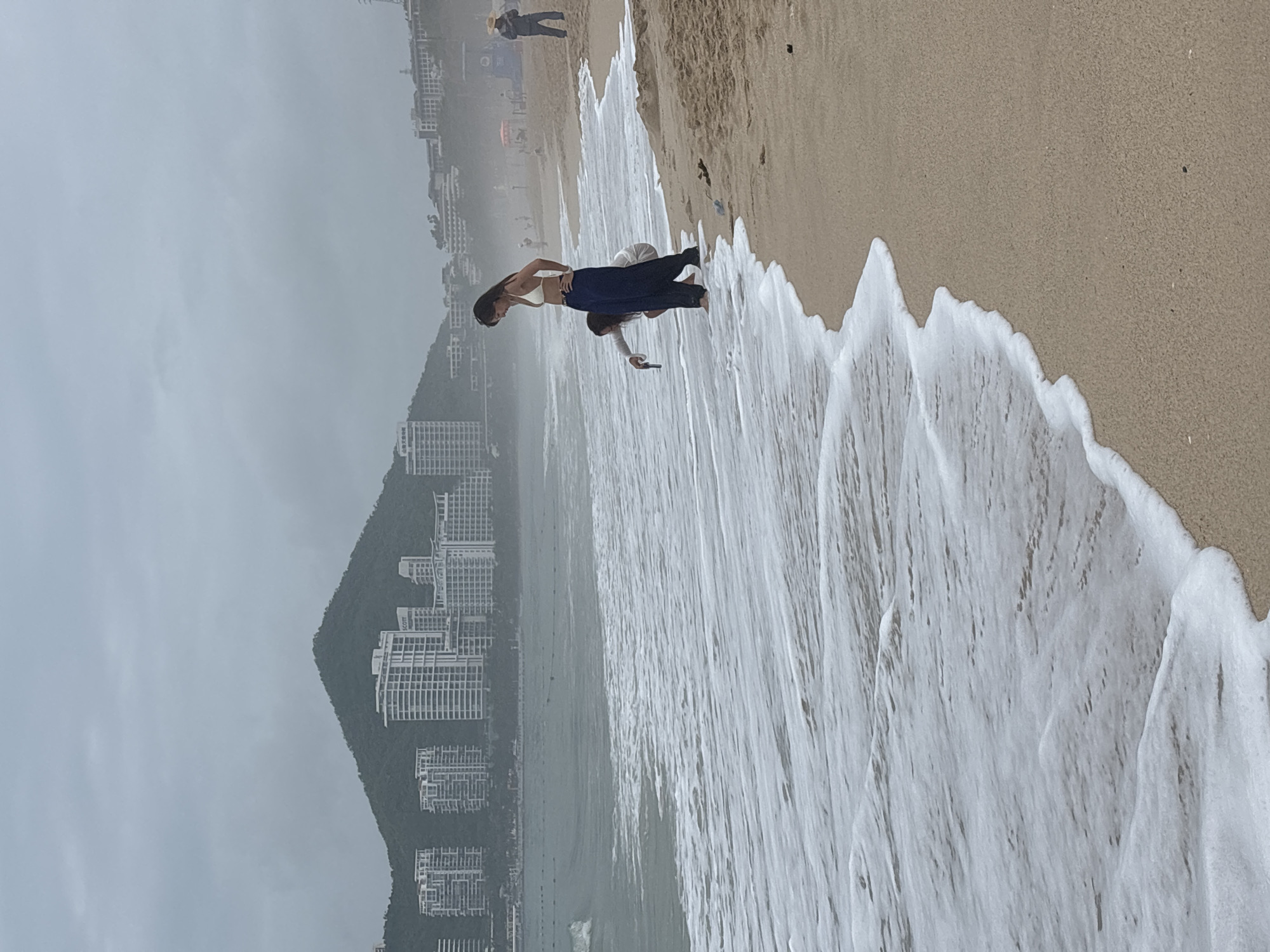 The image depicts a beach scene with two individuals near the water's edge. The person on the left is a woman wearing a white bikini top and dark pants, standing and looking down at the water. To her left and slightly in front, a second person, appearing to be a child or smaller adult, is kneeling and reaching towards the water. The background shows a hazy shoreline with numerous modern white buildings and a large, tree-covered mountain. The sky is overcast and gray, contributing to a subdued atmosphere. The foreground is dominated by the sandy beach and the incoming tide, with white foam washing ashore. The image captures a moment of interaction with the sea on a cloudy day, with a backdrop of urban development integrated with nature.