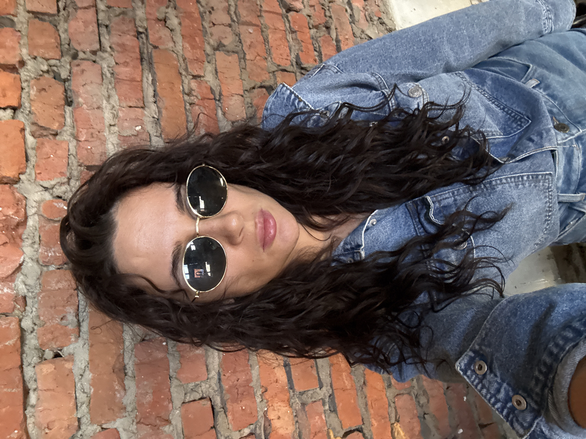 A person with dark, curly hair and sunglasses is lying on their side in front of a brick wall. The person is wearing a blue denim jacket. The location is not discernable, therefore it is not possible to verify it being in Chaikovsky, Russia.