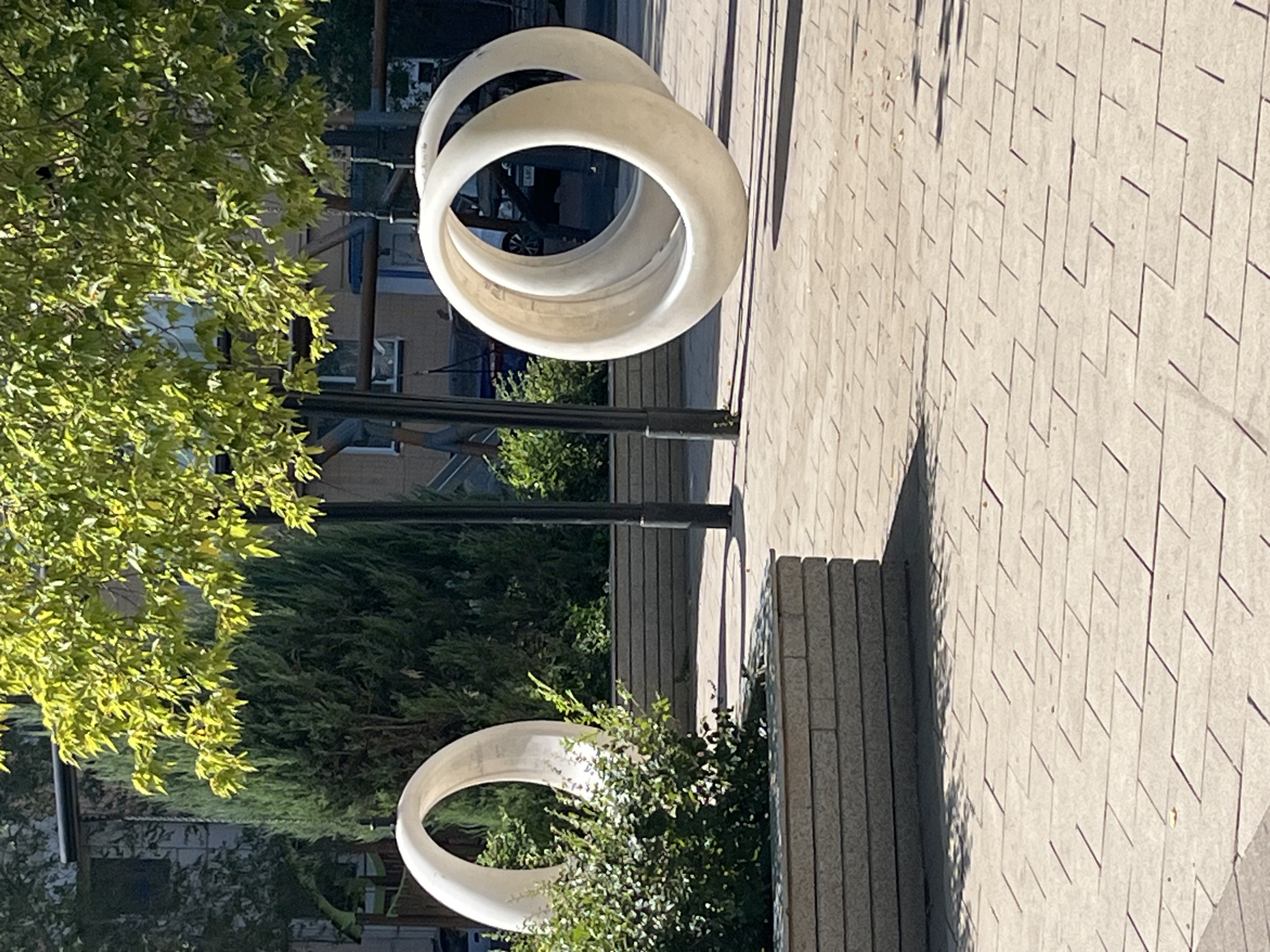 This is an outdoor, eye-level, medium shot of a public space in Astana, Kazakhstan, featuring modern art installations. The foreground is paved with light-colored, interlocking rectangular bricks laid in a staggered pattern, casting shadows from overhead foliage. To the left, a low, rectangular planter box made of stacked grey stone contains a lush green bush. Behind the bush stands a large, smooth, white, ring-shaped sculpture. In the mid-ground, to the right, two similar white ring sculptures are positioned side-by-side, appearing to be slightly tilted. A black lamppost is visible to the right of the rightmost ring sculpture. In the background, behind the sculptures, are mature evergreen trees, including a tall, slender one to the left and broader ones further back, beneath the dappled sunlight filtering through a deciduous tree with vibrant green leaves. Beyond the trees, glimpses of buildings with windows and some outdoor playground equipment are visible. The overall scene depicts a well-maintained urban park or plaza area.