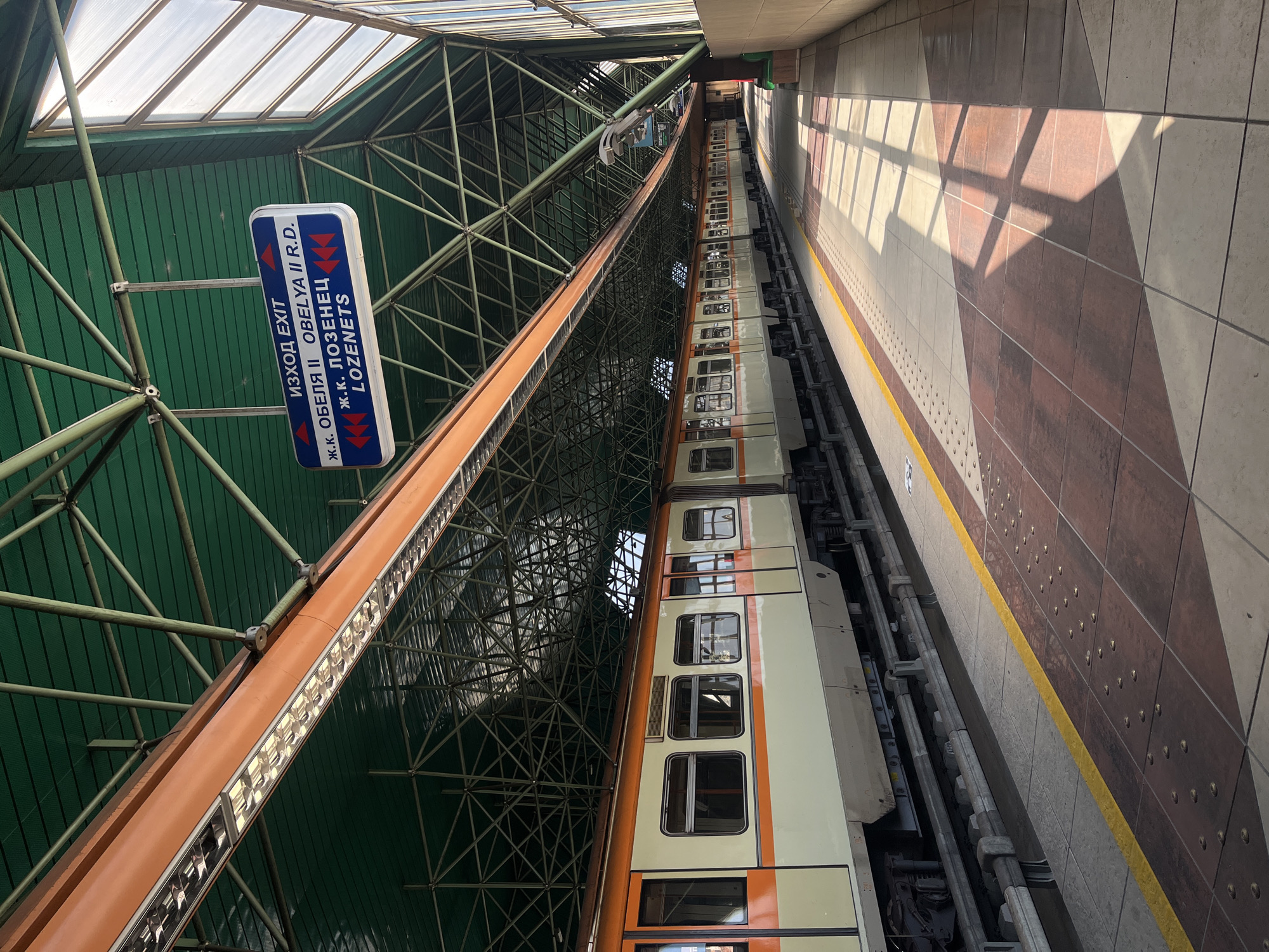 This image depicts a metro station in Sofia, Bulgaria. A beige and orange train is stopped at a platform. Above the train is a metallic structure with a green roof and skylights. A sign is hanging from the structure, with text in Cyrillic and Roman letters indicating "Exit" and the names "Obelya II R.D." and "Lozenets." The platform wall is tiled in brown and beige, with a yellow safety line.