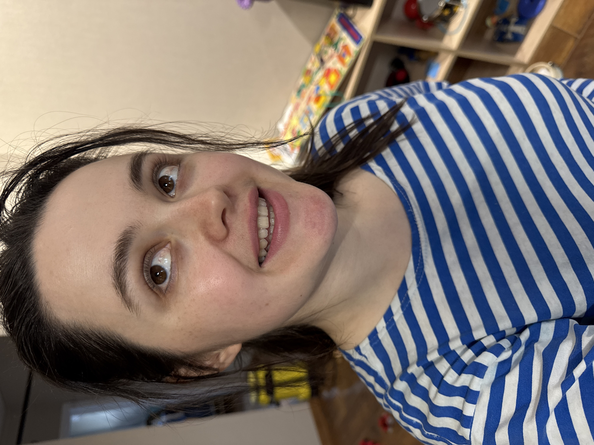 The image is a close-up portrait of a young woman, rotated 90 degrees clockwise, capturing her from a slightly low angle as she looks towards the camera. She has dark hair, dark brown eyes, and fair skin with some freckles. Her eyebrows are dark and well-defined. She is smiling broadly, with her mouth open to reveal her white teeth, conveying a cheerful and engaging expression.
She is wearing a blue and white horizontally striped shirt, a common design reminiscent of a "telnyashka." The background is softly blurred but indicates an indoor setting. Behind her head is a light-colored wall. To her right, a wooden shelving unit is visible, holding various indistinct, colorful objects, possibly toys or decorative items. A portion of a wooden floor can be seen in the lower right. The image captures a personal moment, likely a selfie, within an indoor environment in Izhevsk, Russia. No text is visible in the image.