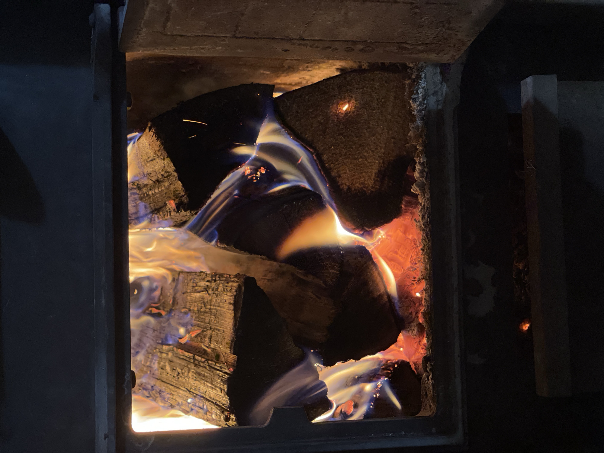 The image depicts a wood-burning stove with logs inside, actively burning. The logs are arranged within the stove's firebox, and flames are visible engulfing them. The interior of the stove is lined with a refractory material. The stove door is slightly ajar. The image gives no evidence as to location in the city of Перевозное, Russia.