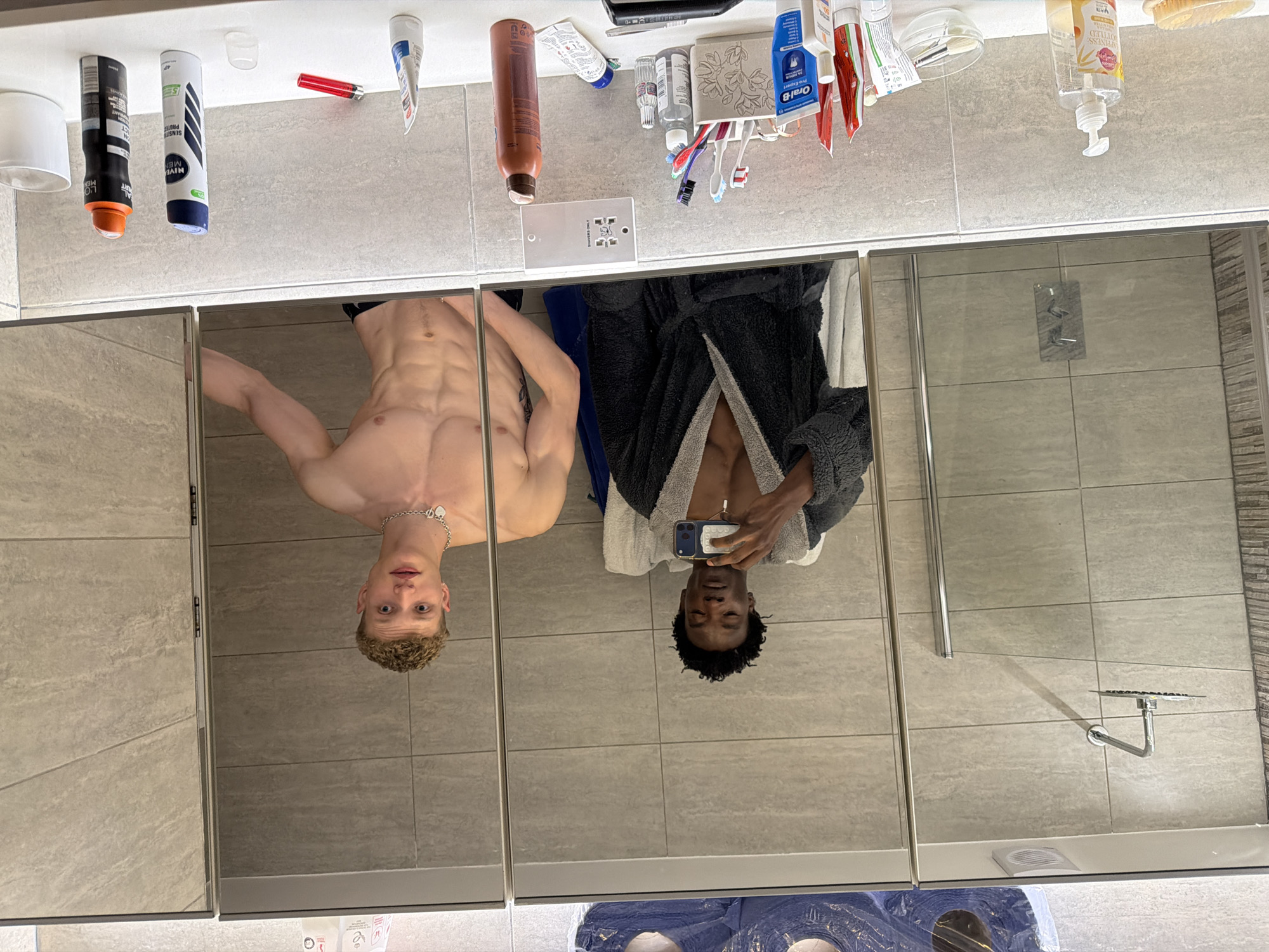 The image displays two young males reflected in a modern bathroom mirror. The setting is located within the United Kingdom, identifiable by the distinct white UK shaver socket present on the tiled wall above the counter. The male on the left, with dark, curly hair, is Black and wears an open dark grey bathrobe, exposing his bare chest with a silver pendant necklace. He holds a smartphone, angled to capture the reflection. The male on the right, with short, curly blonde hair, is White, shirtless, showcasing a muscular torso, and wears dark bottoms. He has a silver chain with a heart-shaped charm and a tattoo on his upper left arm. The bathroom features light grey tiled walls, a glass shower enclosure with a rain shower head, and a white vanity counter. The counter is organized with various toiletries, including a "HAPPINESS BOTTLED" hand soap dispenser, multiple toothbrushes in a white holder, Oral-B toothpaste, Nivea Men and L'Oréal Men Expert deodorant sprays, a red lighter, and a white ceramic mug. A ventilation fan and blue plastic material are reflected along the top edge of the mirror.
