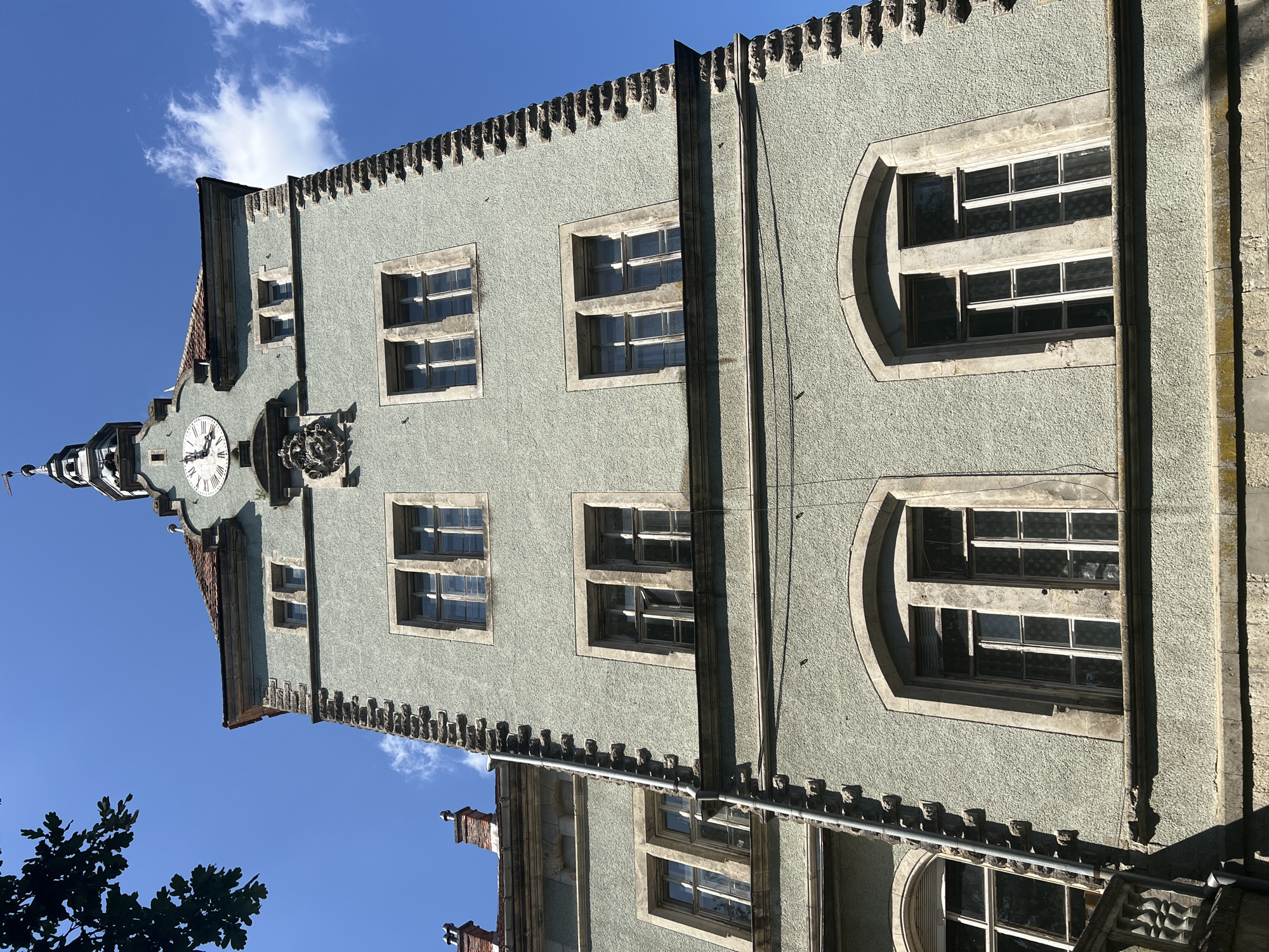The image shows a detailed view of the facade of a light green, multi-story building, possibly a historic structure, in the city of Karpaty, Ukraine. The building features a tower-like structure with a prominent clock face, Roman numerals, and a weather vane at its apex. The facade is characterized by rough plaster, contrasting stone accents along the corners, and multiple windows.
The windows vary in design, with some being rectangular and divided into multiple panes, while others are arched. The architectural style suggests a historical period, with ornate detailing visible in the stonework and around the windows. A section of a red-tiled roof is visible at the top, along with chimneys. The building appears to be under a clear blue sky with scattered white clouds, and a tree branch with green leaves is visible in the upper left corner, casting a shadow. The perspective is from a low angle, looking upwards at the building. No people are visible in the image.