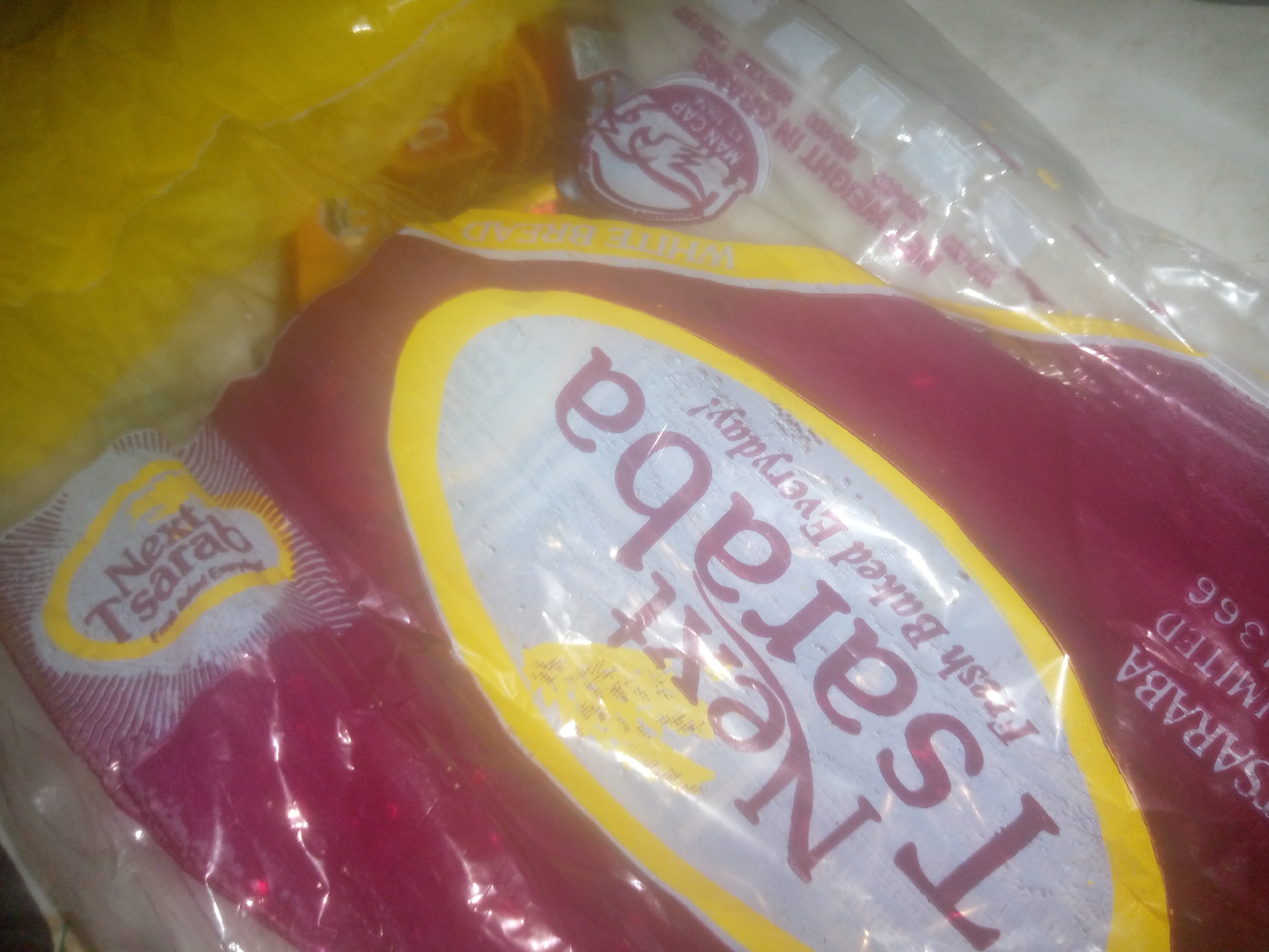 The image shows a close-up of a loaf of bread wrapped in plastic packaging. The bread packaging is primarily red and yellow with white text. The text on the packaging appears to be in English and includes the words "Next Tsarab" and "Fresh Baked Everyday". There is also a circular logo with a bird-like image and the text "MAN CAP EST 1962". The lighting in the image is somewhat dim, and there are reflections on the plastic packaging, suggesting it might be under artificial light. The background is out of focus and appears to be a yellowish, possibly other packaging material. The location is not discernible from the image.