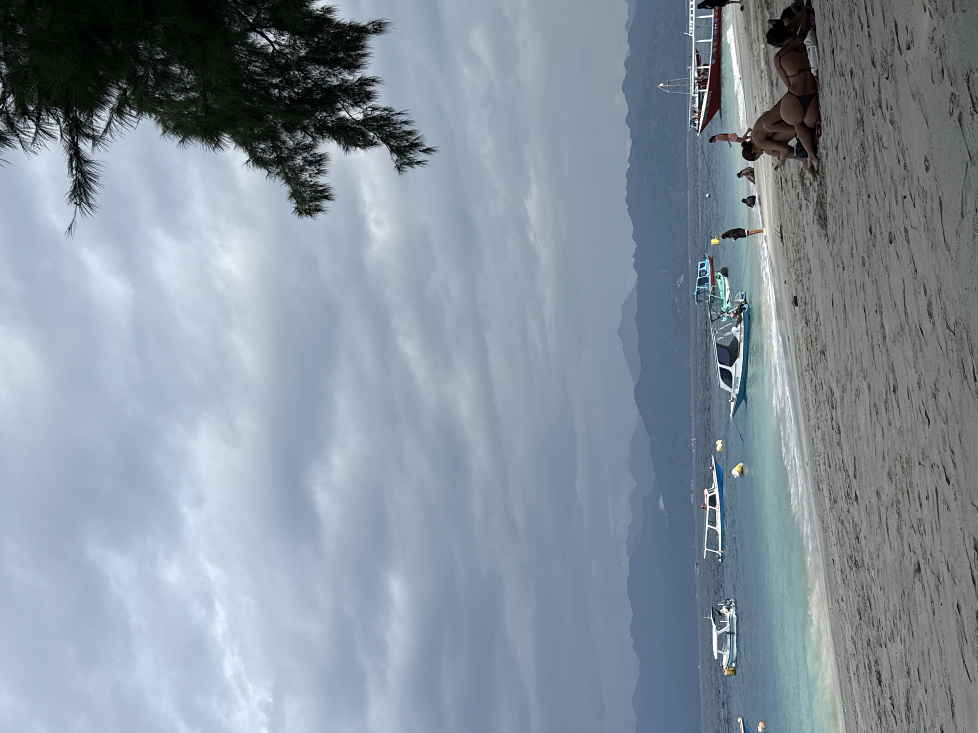 The image depicts a cloudy day at a beach, likely in a tropical location like Indonesia given the presence of boats commonly used for island hopping. The foreground is dominated by a sandy beach where several people are relaxing. Two women are prominently featured in the foreground, lying on towels and appearing to be sunbathing or using their phones. Further back on the beach, other individuals are visible, some standing and others sitting.
The sea is a vibrant turquoise near the shore, transitioning to a deeper blue further out. Several boats are anchored in the water, including smaller speedboats and a larger red vessel with passengers. These boats suggest the location is a popular tourist area, possibly a gateway to other islands.
In the background, a mountain range is visible, shrouded in mist or haze, adding a sense of depth to the scene. The sky is overcast with heavy gray clouds, casting a muted light over the entire landscape. A tree branch with dark green foliage hangs from the upper right corner of the frame, partially obscuring the sky and adding a natural element to the composition. The overall atmosphere is serene, with a slight sense of calm before or after a potential storm, given the cloudy skies.