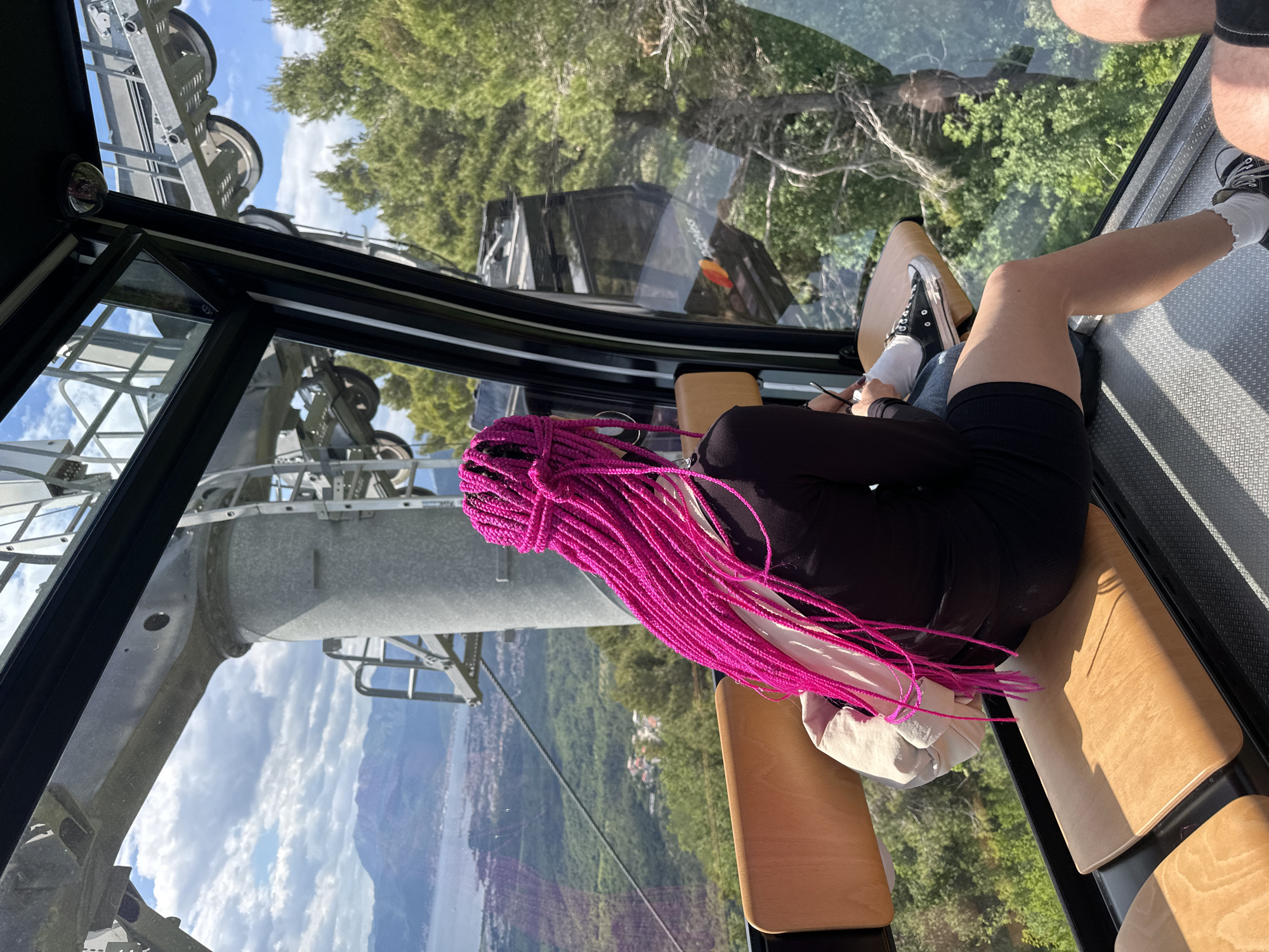 A person with magenta braids sits inside a glass-walled cable car, possibly at the cable car overlooking Čavori, Montenegro. The view outside the glass includes lush green trees, a city landscape, and a body of water. Another person is visible in the frame on the upper right corner. The interior of the cable car includes wooden seats.