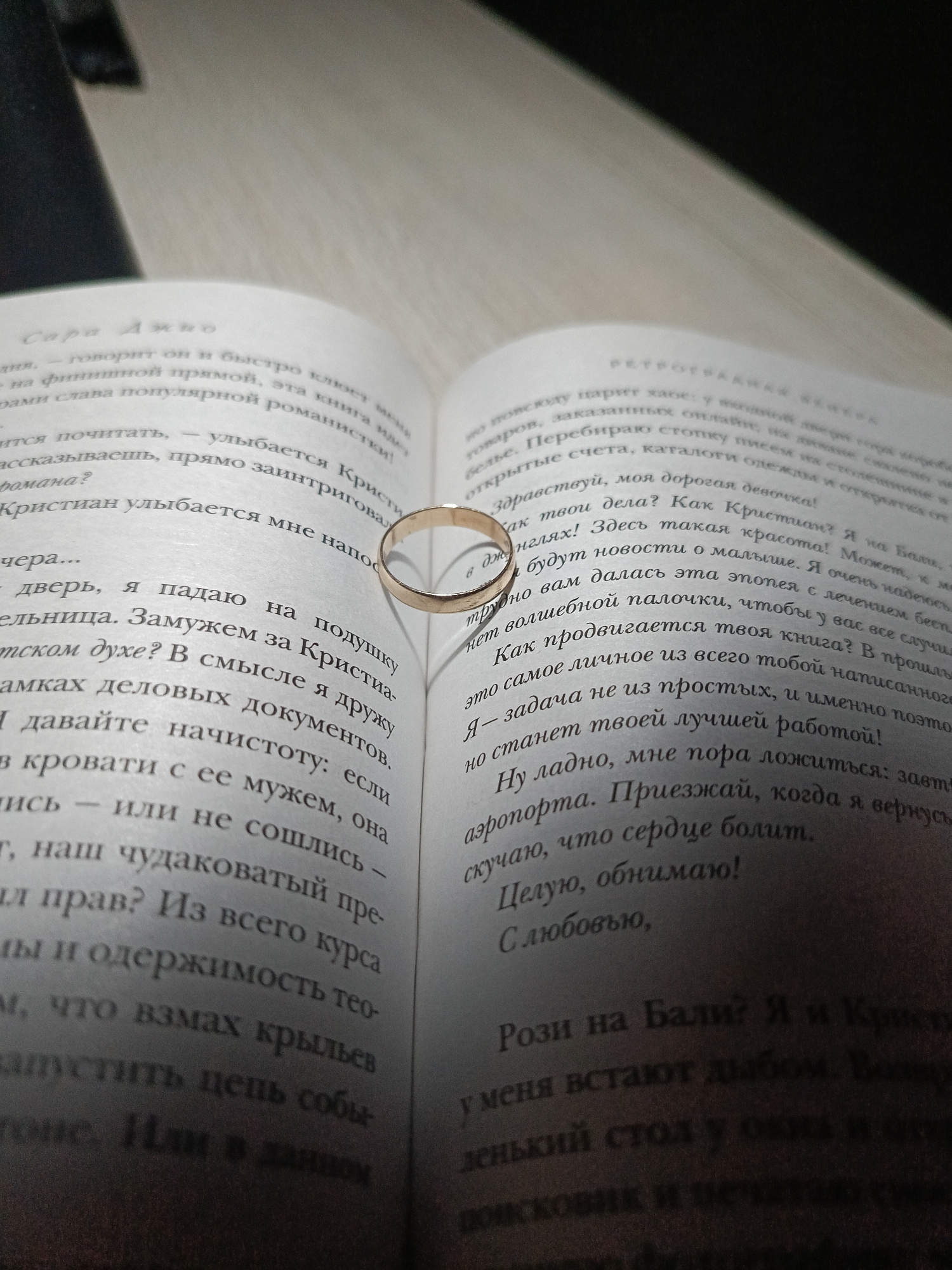 Here is a description of the image:
**Scene:** An open book lies on a light-colored surface, likely a table. A gold ring is placed on the left-hand page of the book, casting a shadow on the page underneath it. The text on the pages is in Russian.
**Objects:** Book, gold ring
**Text Snippets:**
* The left page contains the name "Сара Джио" (Sara Jio) and mentions "Кристиан" (Christian).
* The right page is written in the form of a letter beginning, "Здравствуй, моя дорогая девочка!" (Hello, my dear girl!)
**Environment:** The environment appears to be indoors.
There is no information in the image to indicate the location in Chaikovsky, Russia.