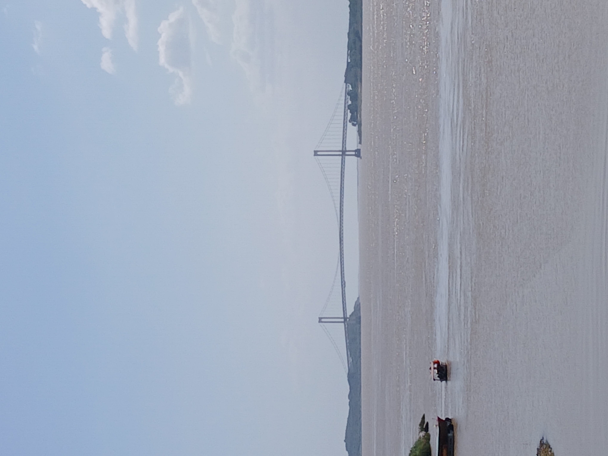 The image shows the Angostura Bridge spanning the Orinoco River in Ciudad Bolívar, Venezuela. The bridge is a suspension bridge with two prominent towers. The river appears wide and muddy. Several small boats or barges are visible near the foreground. The sky is overcast with some scattered clouds. Some shoreline with vegetation can be seen along the riverbanks.