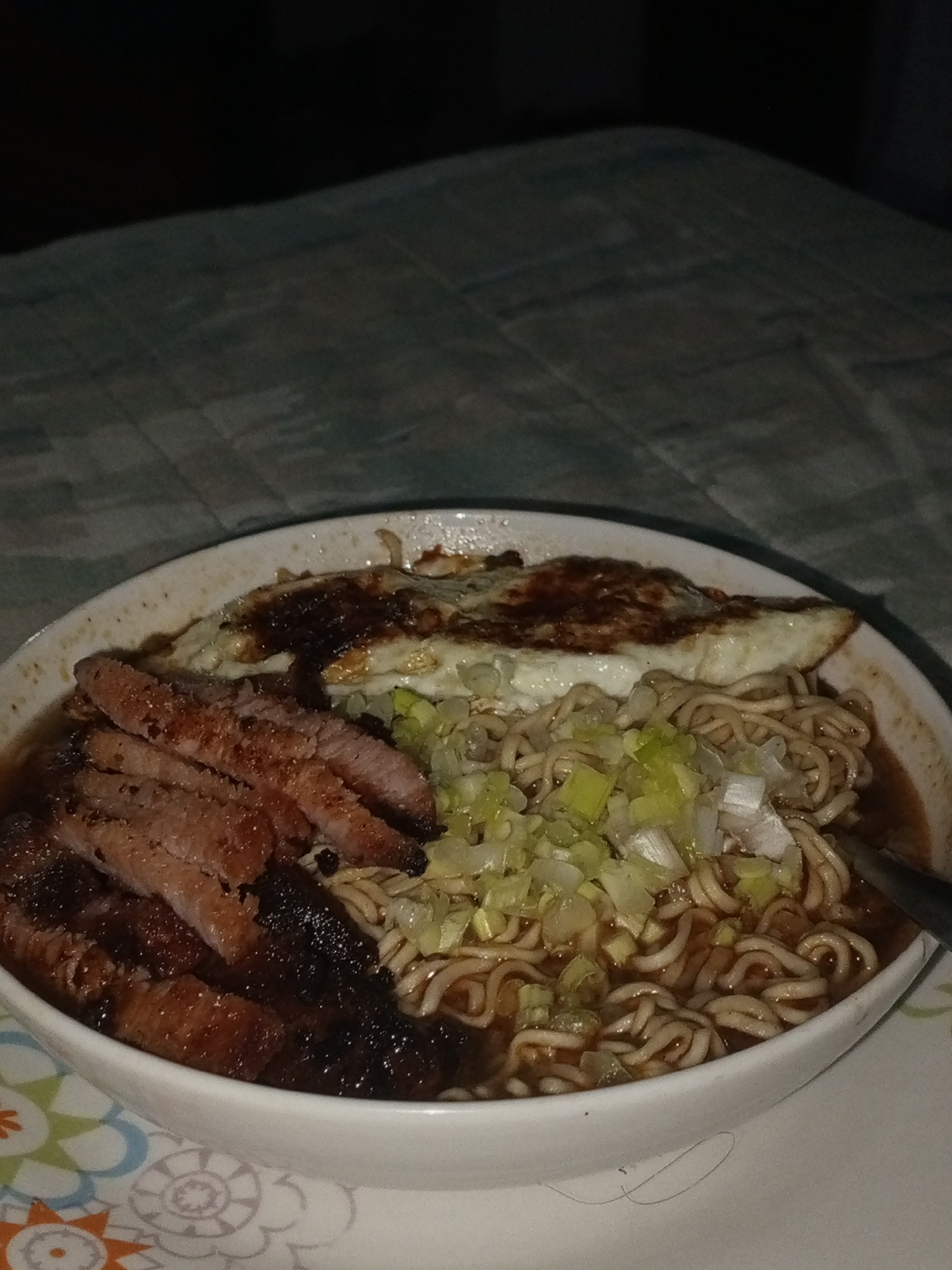A close-up, overhead shot captures a bowl of instant noodles, likely ramen, prepared with various toppings. The white bowl contains a dark broth and curly noodles. Resting on the noodles are several slices of dark, grilled or pan-fried meat, possibly pork or beef, exhibiting some charred areas and a glossy, dark coating. Beside the meat, a fried egg with a crispy, browned top and cooked white is placed. A generous portion of finely chopped green onions (scallions) is scattered over the noodles, adding a fresh green element. A metallic spoon is partially visible submerged in the broth on the right side of the bowl.
The bowl sits on a decorative plate or saucer, partially visible at the bottom of the frame, featuring a colorful floral and geometric pattern in hues of orange, green, and blue. The table beneath is covered with a patterned, dark green or grey tablecloth, which appears textured or checkered. The background is predominantly dark, indicating a dimly lit indoor environment, possibly nighttime. The lighting source appears to be focused on the food, making it the central highlight. There is no visible text in the image. The scene depicts a prepared meal in a casual, domestic setting, consistent with a home in Porlamar, Venezuela.