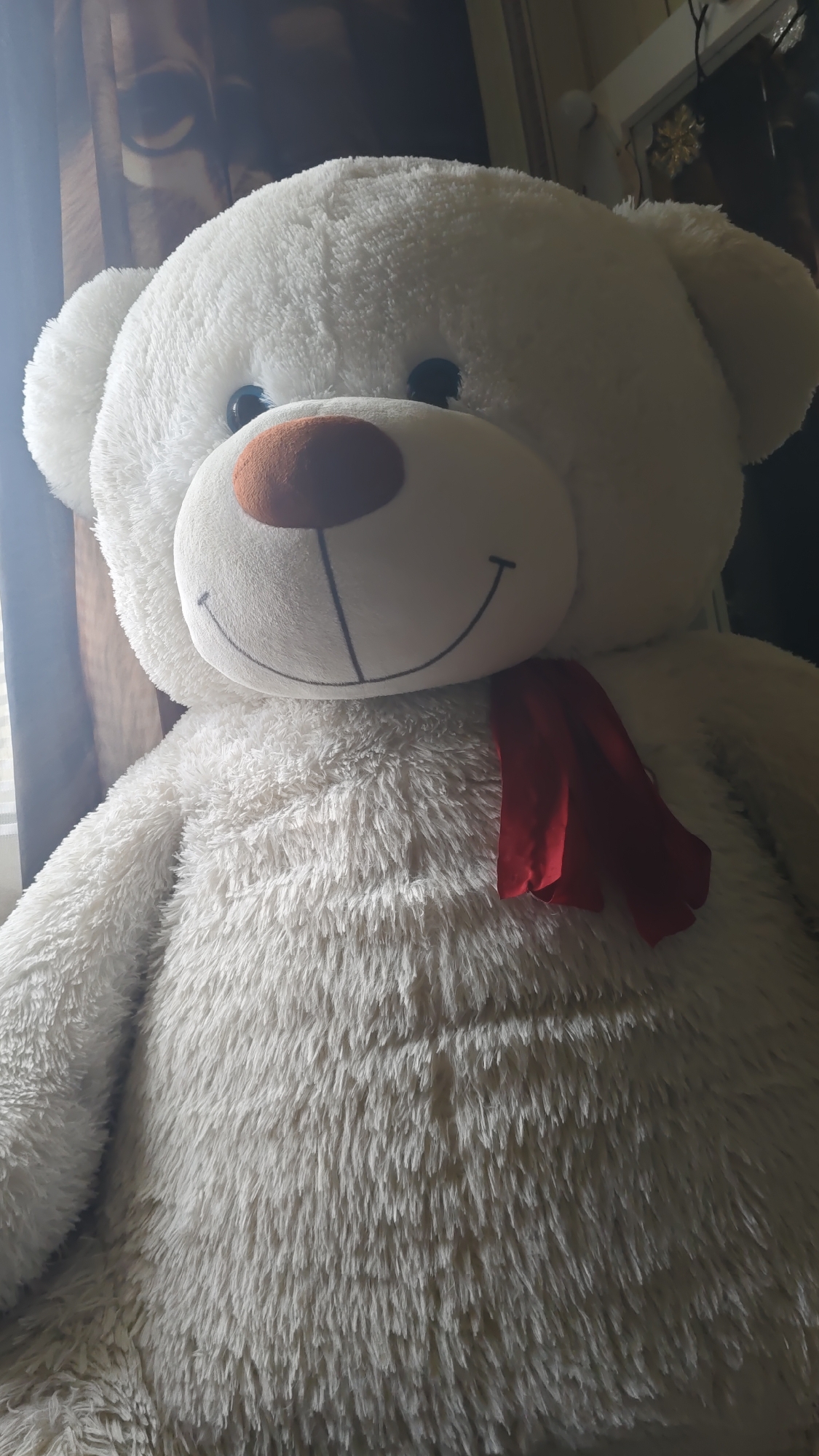 An extreme close-up shot features a large, white, plush teddy bear with a reddish-brown nose and a stitched smile. The bear is wearing a red scarf draped around its neck. The background is dimly lit, with a blurred image of what appears to be a person's face visible through a window or curtain on the left side. The overall lighting suggests an indoor setting, likely a home. No specific details about the city of Karabash, Russia, are discernible from the image.