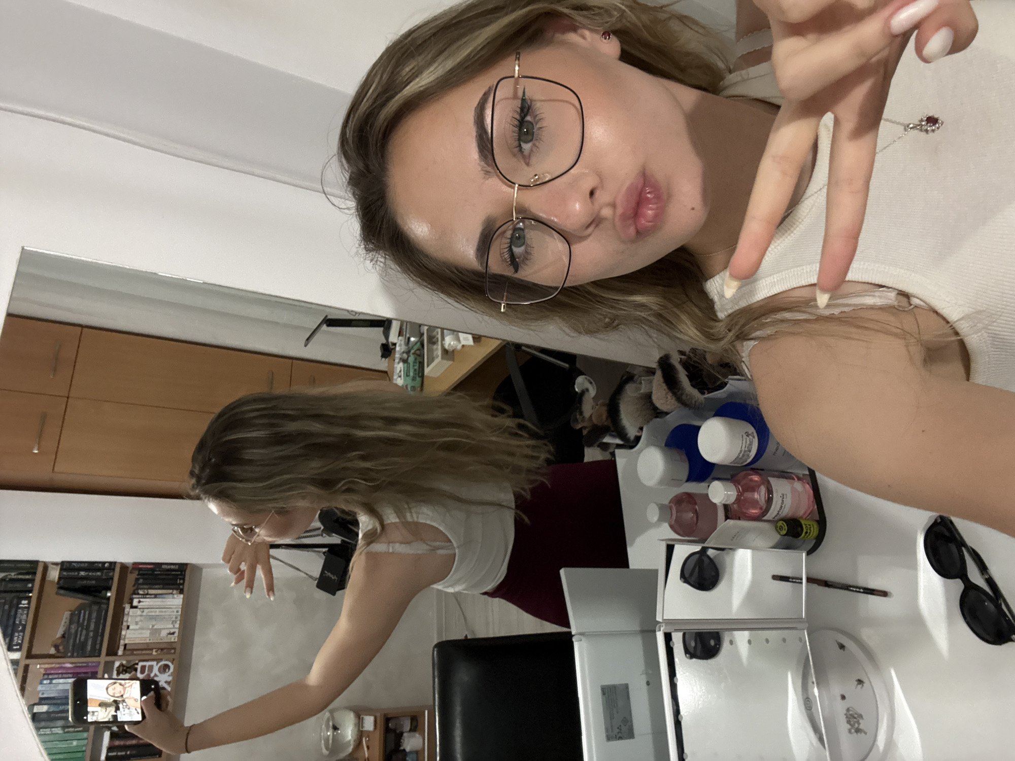 A young woman with blonde hair and glasses is taking a selfie. She's pouting and making a peace sign. Her reflection is visible in a mirror, showing her holding a phone. The background includes a bookshelf, a vanity with makeup, and bottles. The location is not discernible from the image to determine if it is in Blagoevgrad, Bulgaria.