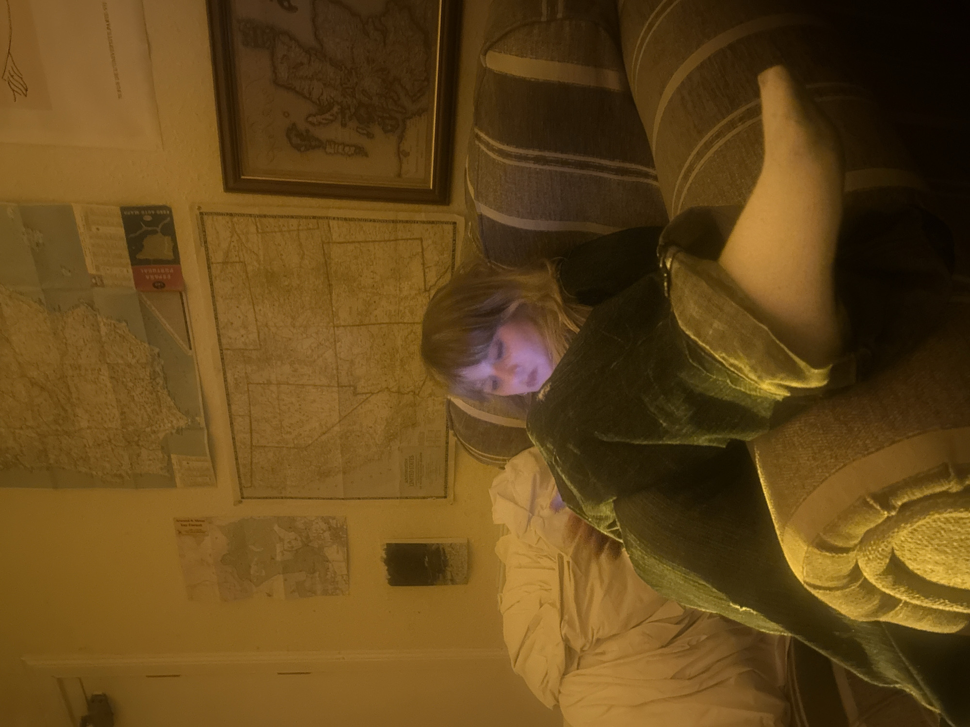 The image captures an indoor scene, likely a living room or bedroom, characterized by warm, dim, yellowish lighting, suggesting evening or nighttime. A young individual with light-colored hair is the central subject, reclining on a striped sofa. Their face is illuminated by the glow of a screen device, indicating they are engrossed in it. The person wears dark trousers or jeans, and one foot, clad in a white sock, is casually propped up on the sofa. A rumpled white blanket or duvet is visible beneath them.
The wall behind the person is decorated with several maps. A large, framed, antique-style map, possibly of Europe, hangs in the upper right. Below it, a large unframed map, partially revealing the text "UNITED STATES," is pinned to the wall. Adjacent to this, a smaller, folded map or booklet is visible, clearly displaying "AUTO MAPA" and "PORTUGAL," with "FORMA PORTUGAL" also decipherable. Another small map is visible lower on the wall, along with a small, dark rectangular object, possibly a speaker or decorative item on a shelf.
The scene depicts a moment of quiet relaxation, with the person focused on their device amidst a collection of geographical wall decor, implying an interest in travel or cartography. The visible text explicitly mentions "PORTUGAL" and "UNITED STATES."