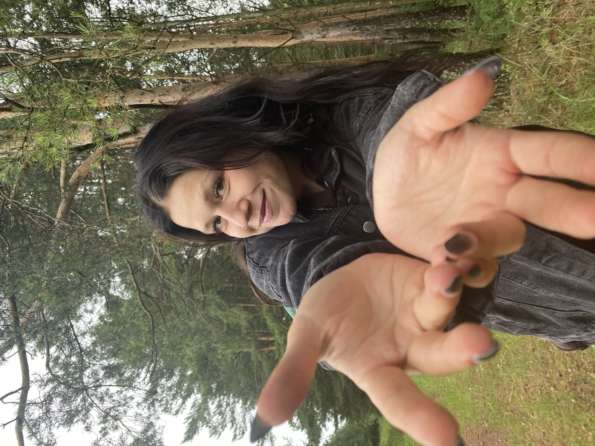 This is a vertical, eye-level, close-up shot of a young woman in a forest. She has long, dark, wavy hair and is wearing a dark denim jacket. Her face is turned towards the camera, and she is smiling. Her hands are outstretched towards the camera, with her palms facing outwards. Her fingernails are painted a dark gray color.
The setting appears to be a pine forest, with tall trees and green grass visible in the background. The lighting suggests it is daytime, possibly overcast. The overall emotion conveyed is friendly and inviting. There is no visible text in the image.