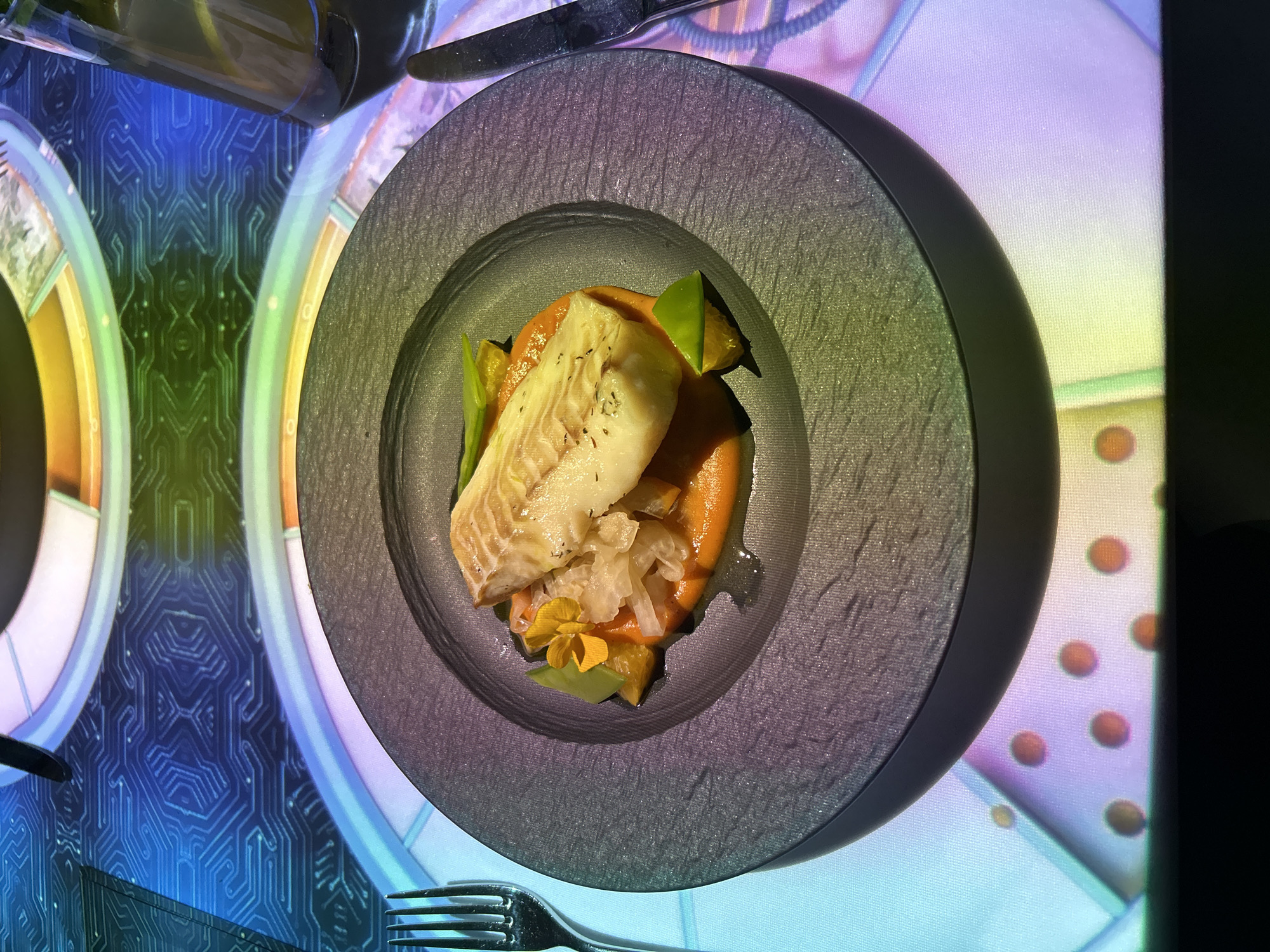A top-down shot shows a plate of food on a table with a digitally projected surface. The food on the plate appears to be a piece of white fish, possibly cod or halibut, resting on a bed of orange puree, garnished with vegetables, and a small yellow flower. The plate is a dark gray, textured, round bowl. Silverware, including a fork and knife, are positioned on either side of the plate. The table's surface is digitally projected with futuristic geometric patterns in shades of blue, yellow, green, and purple. The scene is from a restaurant in Warsaw, Poland.