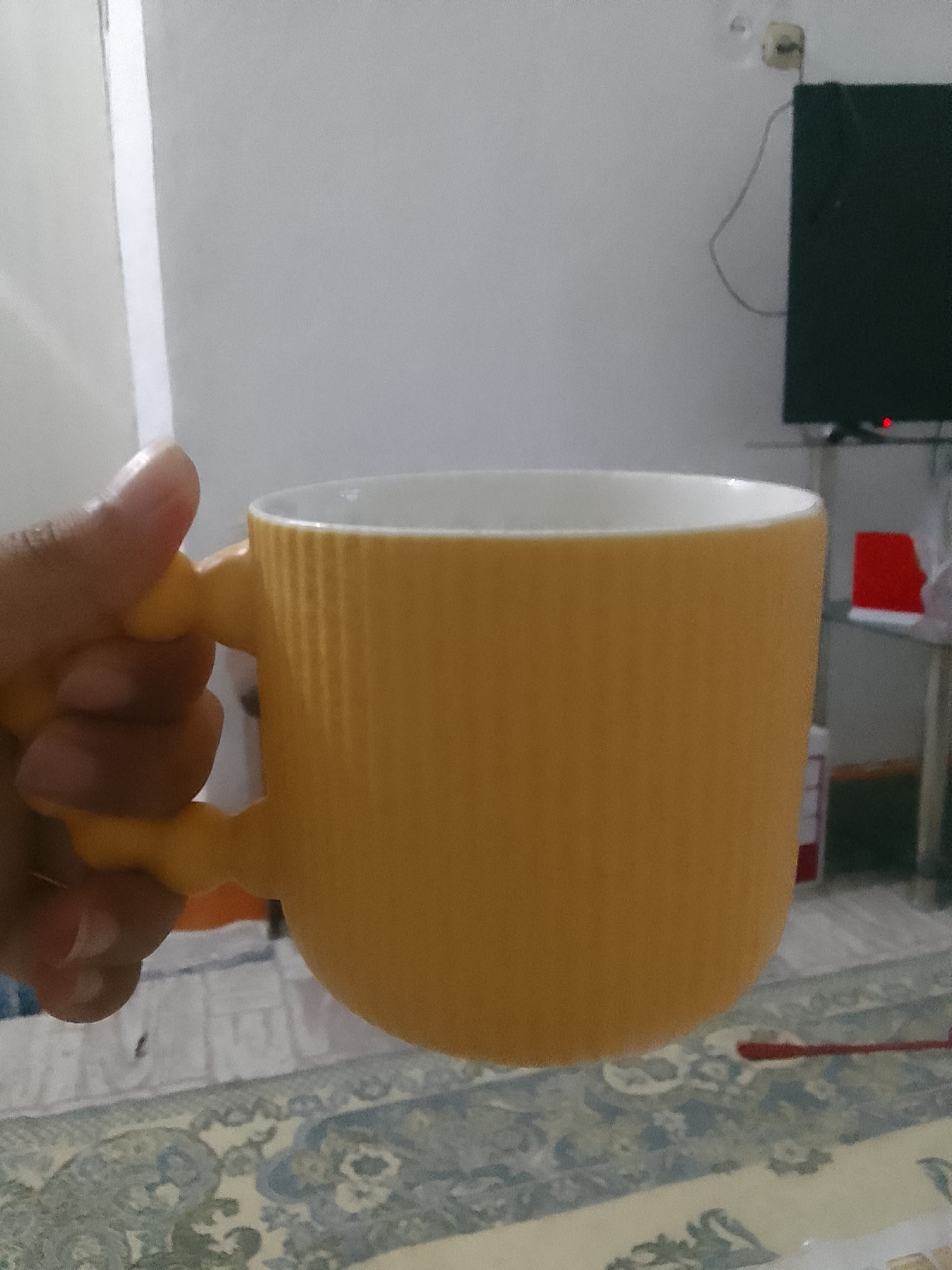 A person's hand is holding a large, ribbed, yellow mug with a white interior. The mug is positioned in the foreground, obscuring a portion of the background. Visible in the background are a dark television set with a red indicator light, a shelf with a red object, and a patterned rug on the floor. The scene appears to be indoors, in a room with neutral-colored walls. There is no indication of the city of Taxtakópir, Uzbekistan in this image.