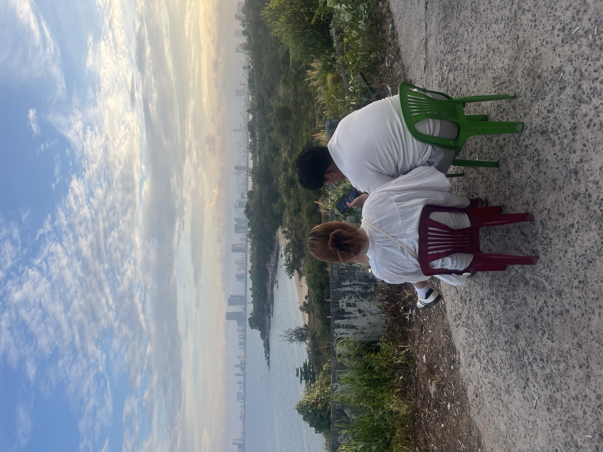 Two individuals, a woman and a man, are seated on plastic chairs facing away from the camera, overlooking a panoramic view of Đà Nẵng, Vietnam. The woman, on the left, is wearing a white top and is seated on a red chair. The man, on the right, is wearing a white polo shirt and grey shorts and is seated on a green chair. Both appear to be engaged with their mobile phones. The foreground features a dirt and gravel path with sparse vegetation and debris. Behind them, a concrete wall separates the elevated viewpoint from a lush, green landscape that slopes down towards a beach and the sea. The city of Đà Nẵng, with its numerous high-rise buildings, is visible in the distance across the water, under a sky filled with a mixture of white and grey clouds, with hints of yellow and pink suggesting either sunrise or sunset. The overall scene depicts a relaxed moment of observation and personal engagement with technology against the backdrop of a coastal cityscape.