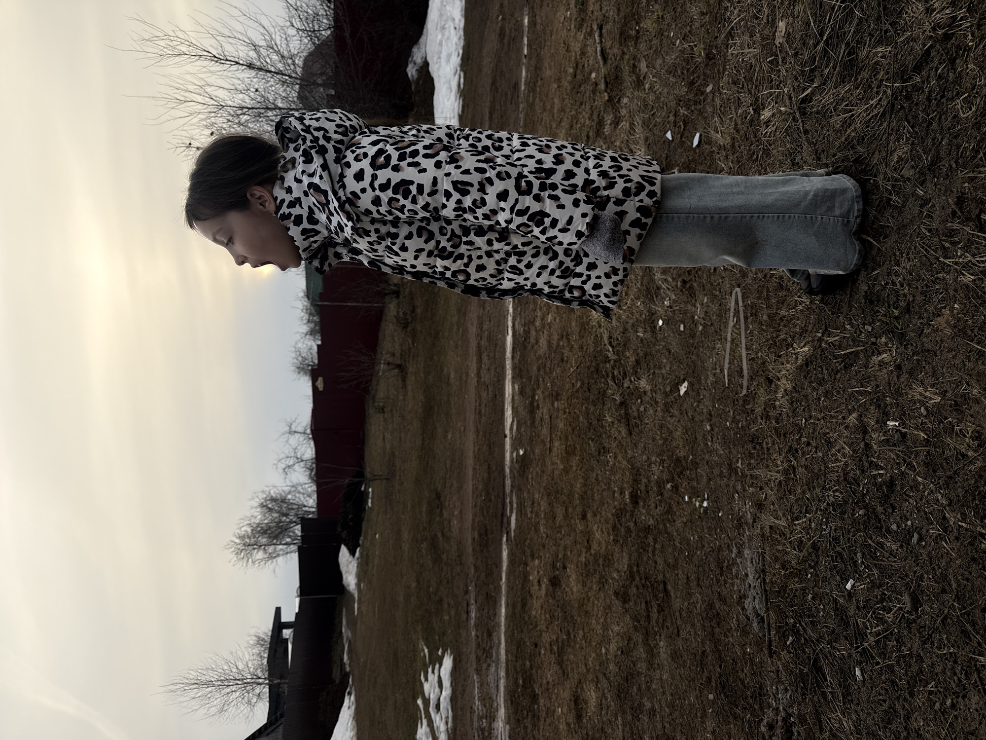 A person stands in a grassy yard in Votkinsk, Russia. They wear a spotted coat and jeans. The ground is covered in brown grass and some snow. A fence and building are in the background. The sky is overcast.