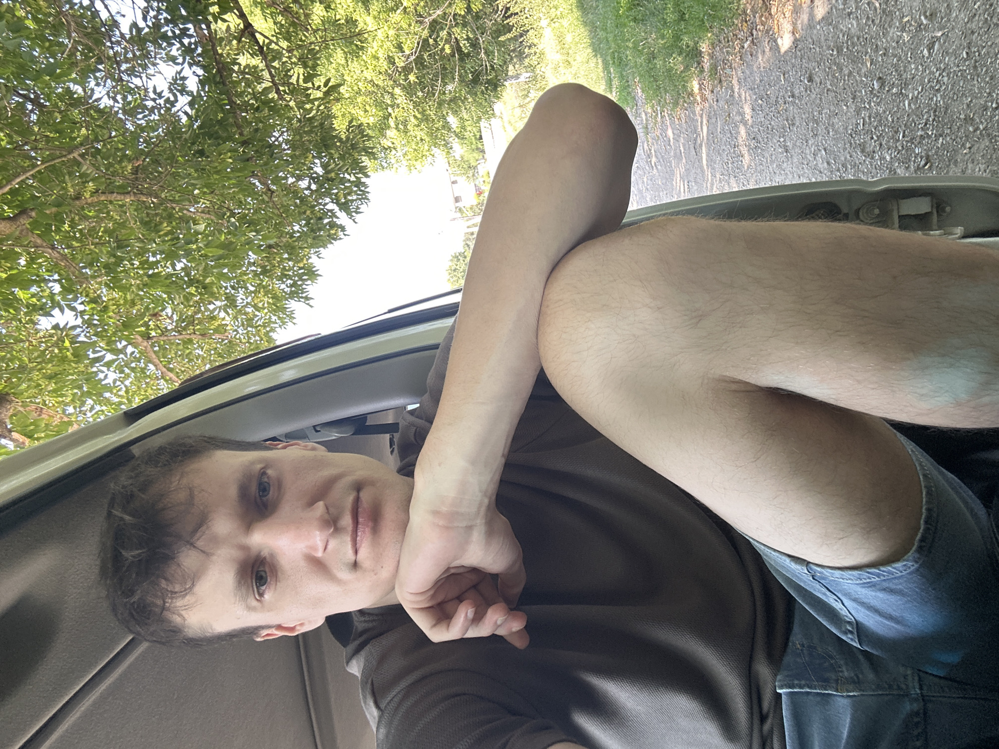 A young Caucasian male is seated in the open door of a vehicle, his body angled towards the camera. He is wearing a dark brown t-shirt and denim shorts. His left arm is resting on his bent knee, and his chin is propped on his hand. The background reveals a sunlit, rural environment with trees and a gravel road. The scene is captured outdoors on a sunny day. The location of Makiv, Ukraine, is not identifiable from the visual information provided in the image.