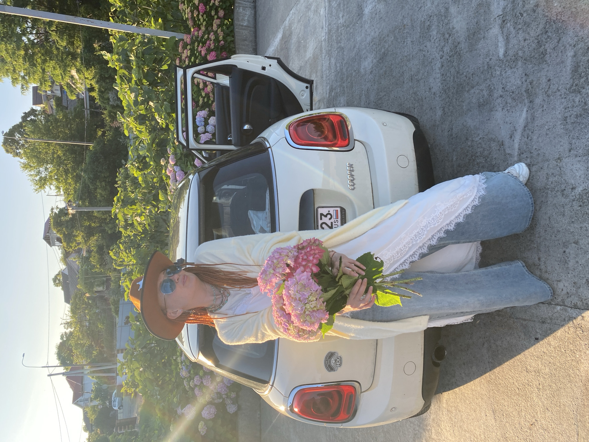 A woman stands next to a white Mini Cooper with the driver's side door open in what appears to be an affluent residential area of Sochi, Russia. The woman wears a brown cowboy hat, large sunglasses, a cream-colored cardigan over a white lace-trimmed dress, and light blue wide-leg jeans. She holds a large bouquet of pink and purple hydrangeas. The Mini Cooper's license plate reads "23 RUSSIA". The scene is bathed in the warm glow of late afternoon sun, casting long shadows. Behind the car, a hillside is densely covered with lush green trees and houses. A line of vibrant pink and purple hydrangeas blooms in front of a low wall. The ground is paved with concrete.