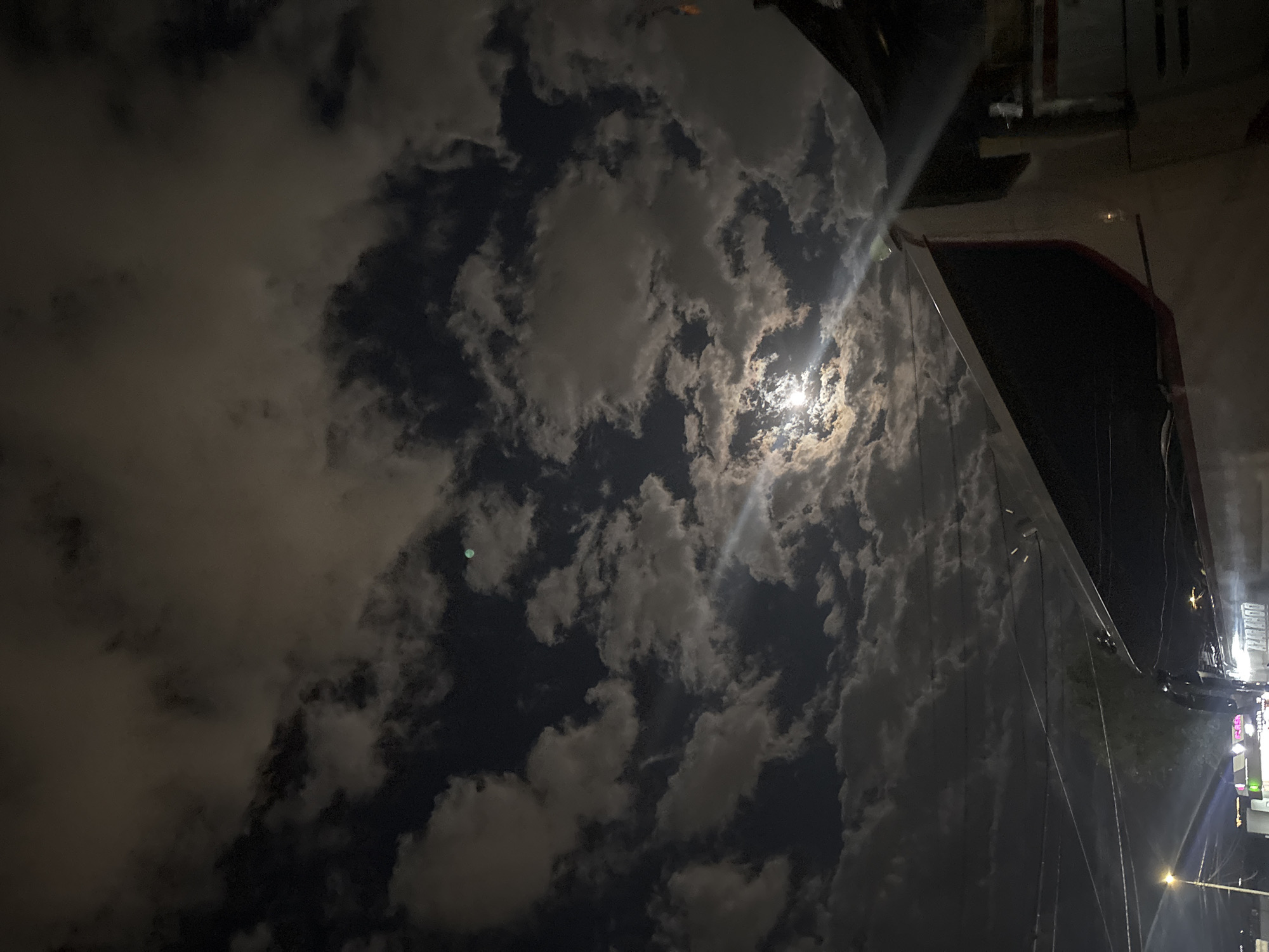 The image depicts a nighttime scene with a sky filled with scattered clouds and a bright moon shining through. The photograph is taken from a low angle, possibly street level, and includes a building structure on the right side. The building appears modern with reflective surfaces. The location may be in the city of Yerevan, Armenia.