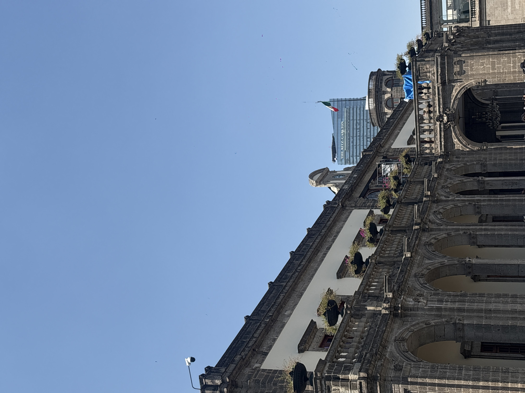This image captures a clear, bright blue sky above a dramatic architectural composition in Mexico City, showcasing a blend of historic and modern structures. The foreground is dominated by a majestic, ornate stone building, likely an ancient cathedral or colonial-era structure, viewed from a low angle. This building features multiple levels with decorative arches, intricate stonework, and balconies adorned with planters containing foliage and pink flowers. A section of the upper facade is draped with a bright blue tarp or fabric, near a rounded, domed part of the building.
In the mid-ground, a towering, modern glass skyscraper stands prominently against the sky. The words "THE RITZ-CARLTON" are clearly visible vertically along its side. A Mexican flag, with its green, white, and red stripes, flies from a pole near the top of this contemporary building. This juxtaposition vividly illustrates the historical depth and modern development of Mexico City.
The sky is a brilliant, cloudless blue, indicating a sunny day, likely late morning or early afternoon. Numerous small, dark specks, probably birds, are visible scattered across the sky. In the lower left corner, a black street light fixture with a camera attached is partially visible. The scene is devoid of people, focusing instead on the architectural landscape and the vibrant sky.