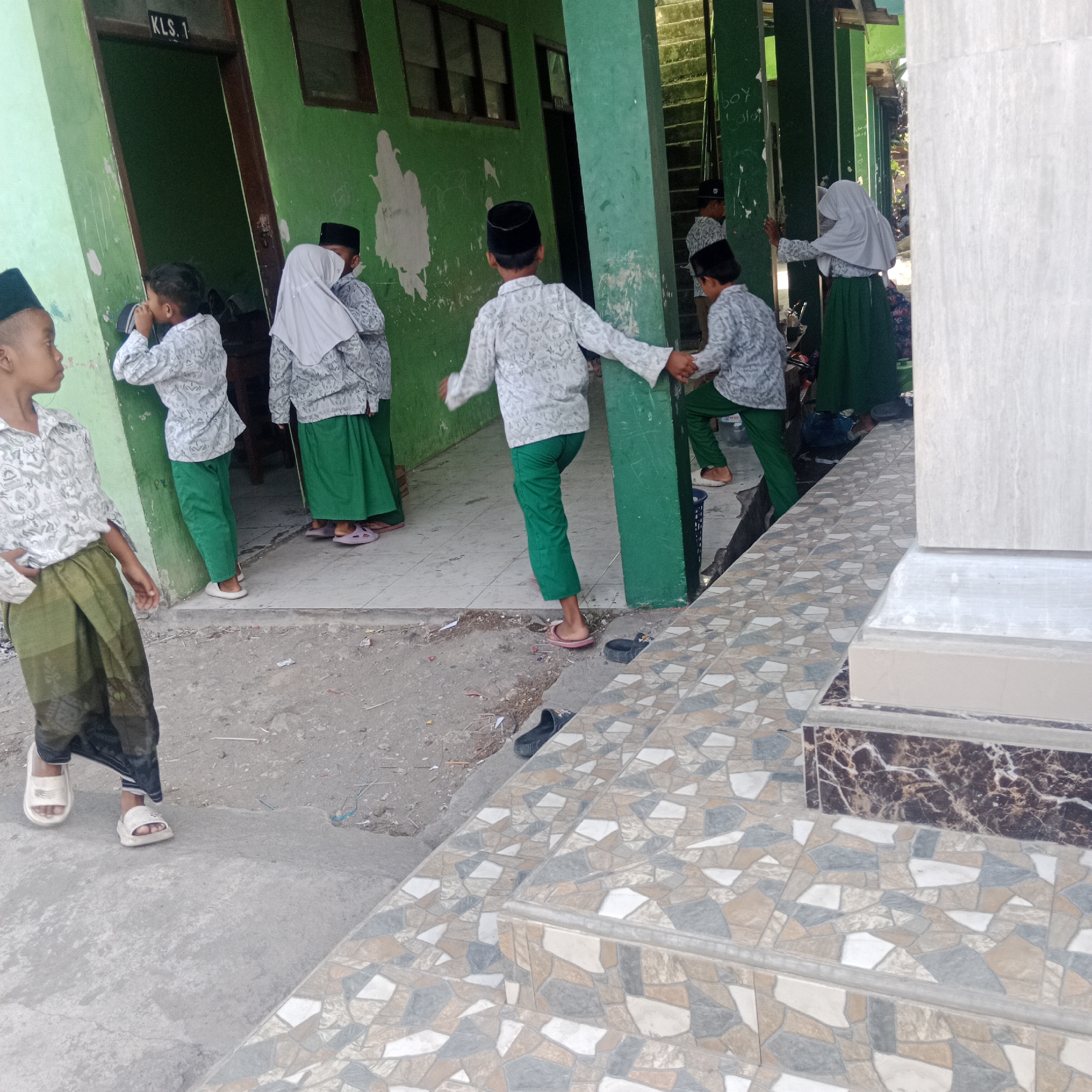 The image depicts a group of children in traditional clothing standing outside what appears to be a school building in Tegowanu Kulon, Indonesia. The building has a green-painted facade with a door labeled "KLS. 1". Several children are wearing white patterned shirts and green pants or skirts, with some also wearing black skullcaps and headscarves. One child in the foreground is facing left, wearing a green sarong-like garment and white sandals. Another child is seen from behind, wearing a white patterned shirt and green pants. In the middle ground, two children are standing near an entrance, one wearing a white patterned shirt and green pants, and another wearing a white patterned shirt and a green skirt with a headscarf. Further back, more children are visible, including one wearing a white patterned shirt and green pants, and another with a headscarf. The environment includes a concrete walkway and steps tiled with a mosaic pattern of brown, beige, grey, and white irregular shapes. The ground is dusty and scattered with debris. The overall scene suggests a casual gathering or break time for the children outside their school.