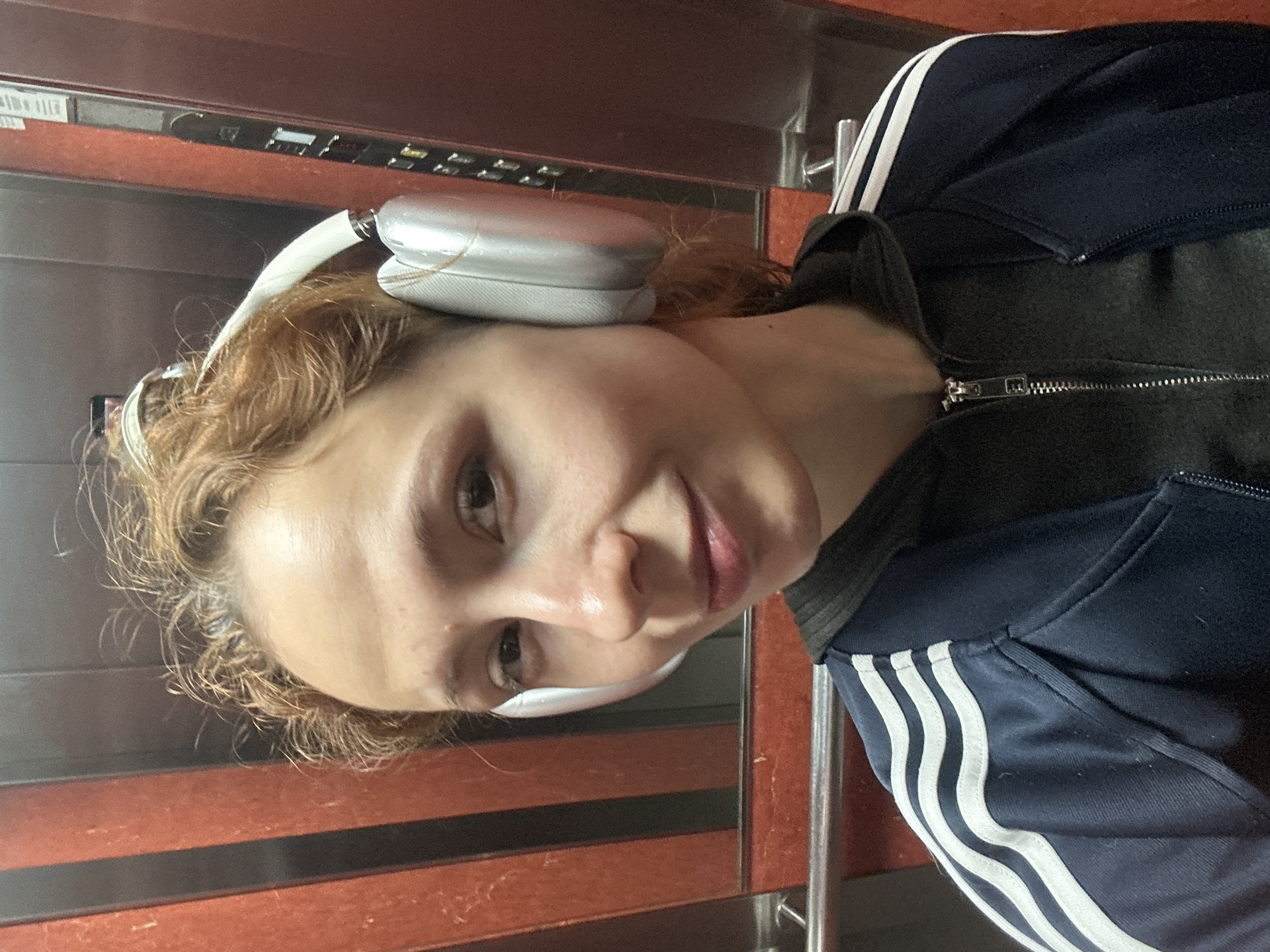 A young woman with reddish-brown, curly hair is depicted in a selfie or close-up portrait, taken from a slightly low angle. She is looking directly at the camera with a calm expression and a faint smile. She is wearing silver over-ear headphones with a white headband. Her attire includes a dark blue tracksuit jacket featuring distinctive white stripes on the sleeves, indicative of an Adidas brand item. A silver zipper is visible on the front of her jacket.
The setting appears to be an elevator interior. The background features metallic panels with a reddish-brown and dark striped pattern. In the upper portion of the image, what seems to be part of an elevator control panel or display is visible, though any potential text or symbols on it are indistinct and unreadable. The overall lighting suggests an indoor environment. The location context provided is Novi Sad, Serbia.