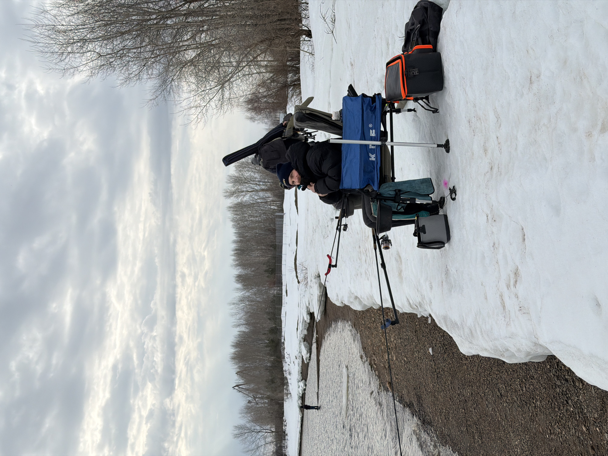 A person is seated on a blue folding chair set up on a patch of snow near a body of water, possibly a river or lake, in what appears to be the outskirts of a city, likely Chaikovsky, Russia, given the context. The person is wearing a dark jacket and a dark-colored hat. They are surrounded by fishing equipment, including fishing rods mounted on supports. Various bags and containers are placed around the chair, potentially holding fishing gear. The ground is covered with snow transitioning to a mix of snow and dirt near the water's edge. In the distance, there are trees with bare branches, suggesting it is late winter or early spring. The sky is overcast with a variety of clouds. A figure can be seen further out in the water.