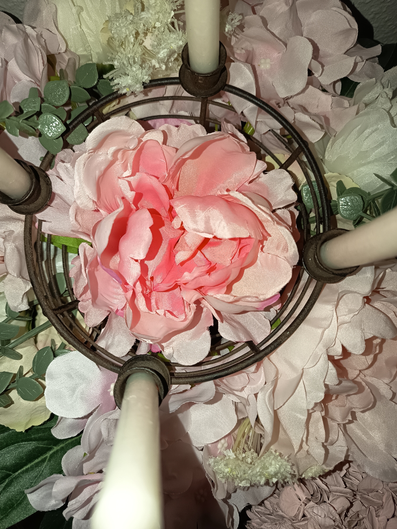 The image shows a close-up, overhead view of a floral centerpiece with candles. A pink peony dominates the center, encircled by a rusty metal ring with four candle holders. White candles are placed in the holders. The peony is surrounded by other pink and white flowers and greenery. The location in Wangen bei Olten, Switzerland, is not discernible from the image.