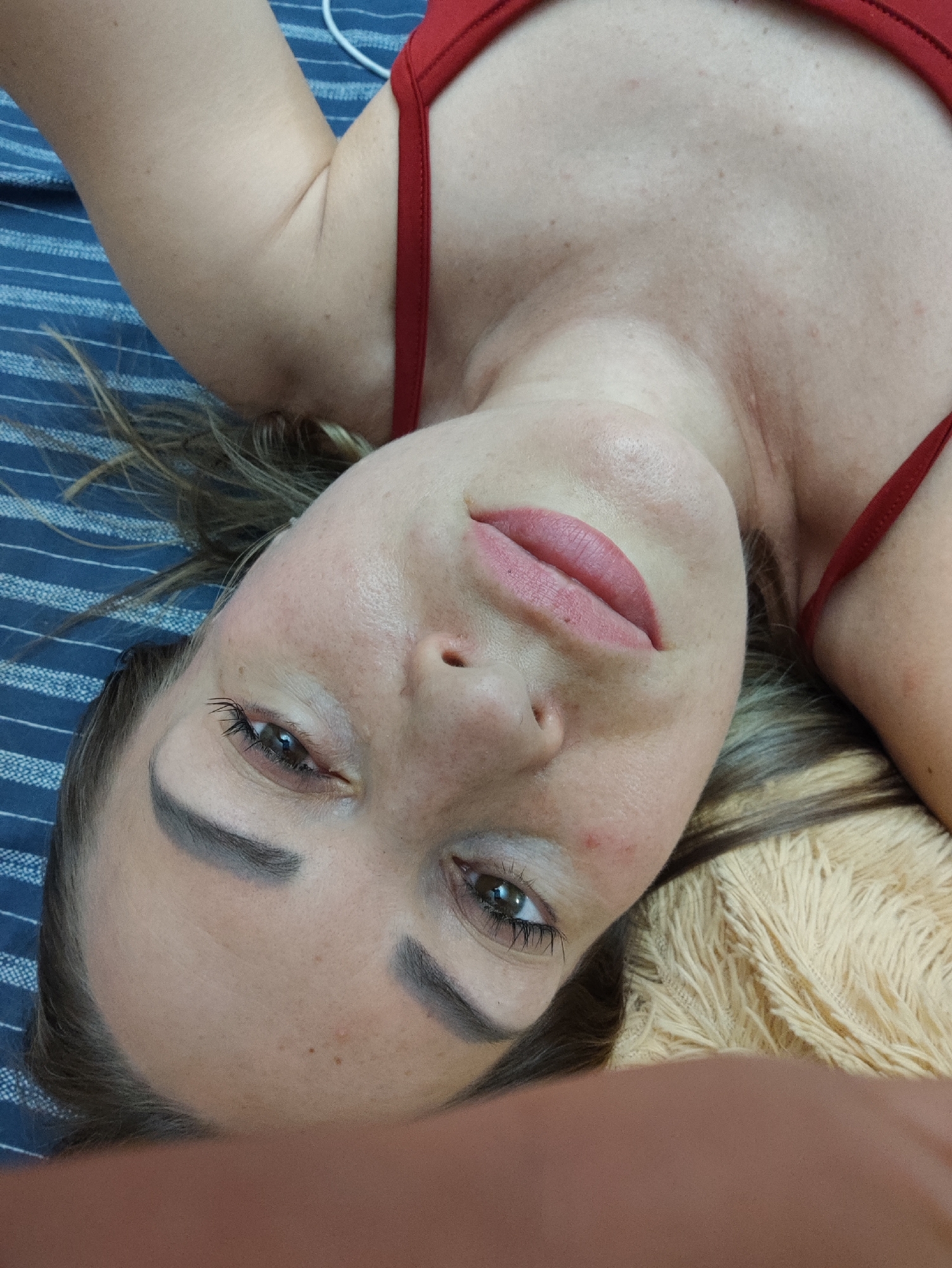 The image is a close-up, tilted selfie of a woman lying on her back. She is wearing a red bikini top. Her face is the central focus, with her head turned to her left and her eyes looking upwards. Her lips are painted a light pink. The background consists of a blue and white striped surface, possibly a bedspread or towel, and a furry, cream-colored blanket or pillow. The lighting suggests an indoor setting. There is no discernible location in the city of Almaty, Kazakhstan, visible in the image.